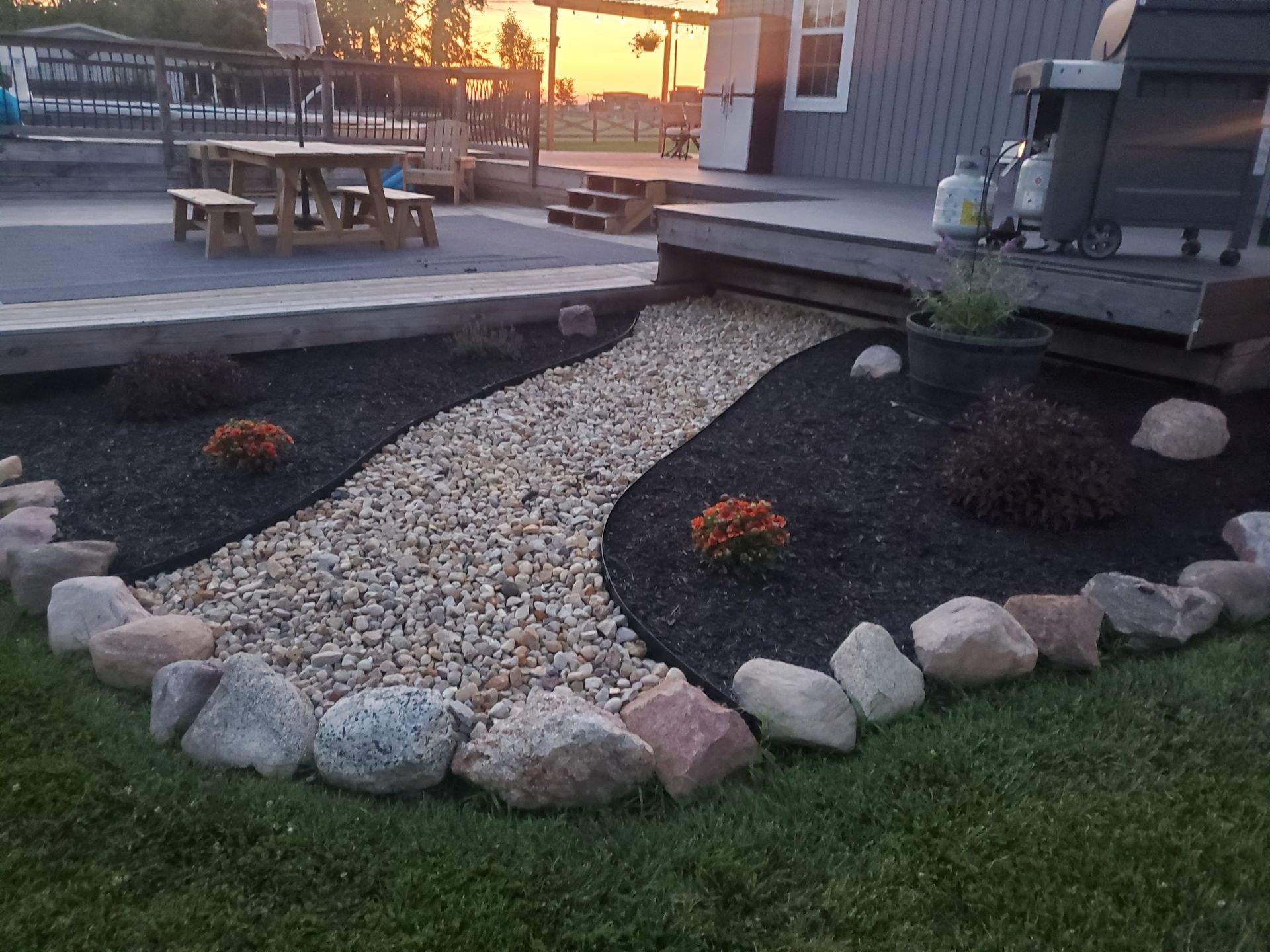 Stone-bordered garden bed with mulch and river of light gravel; near a deck with a grill and picnic table at sunset.