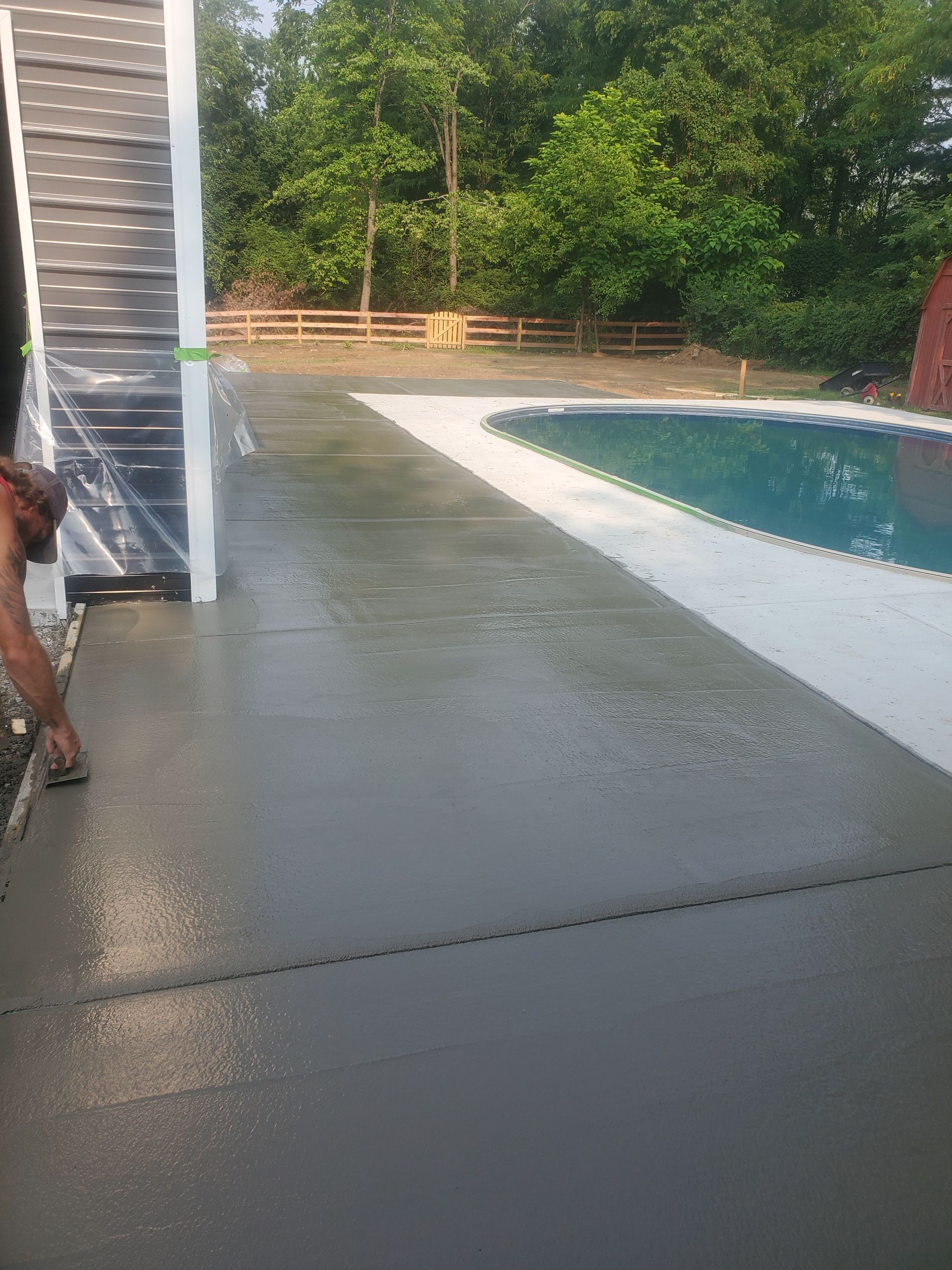 Freshly poured concrete patio next to a swimming pool. A person is working the edge. Trees in the background.