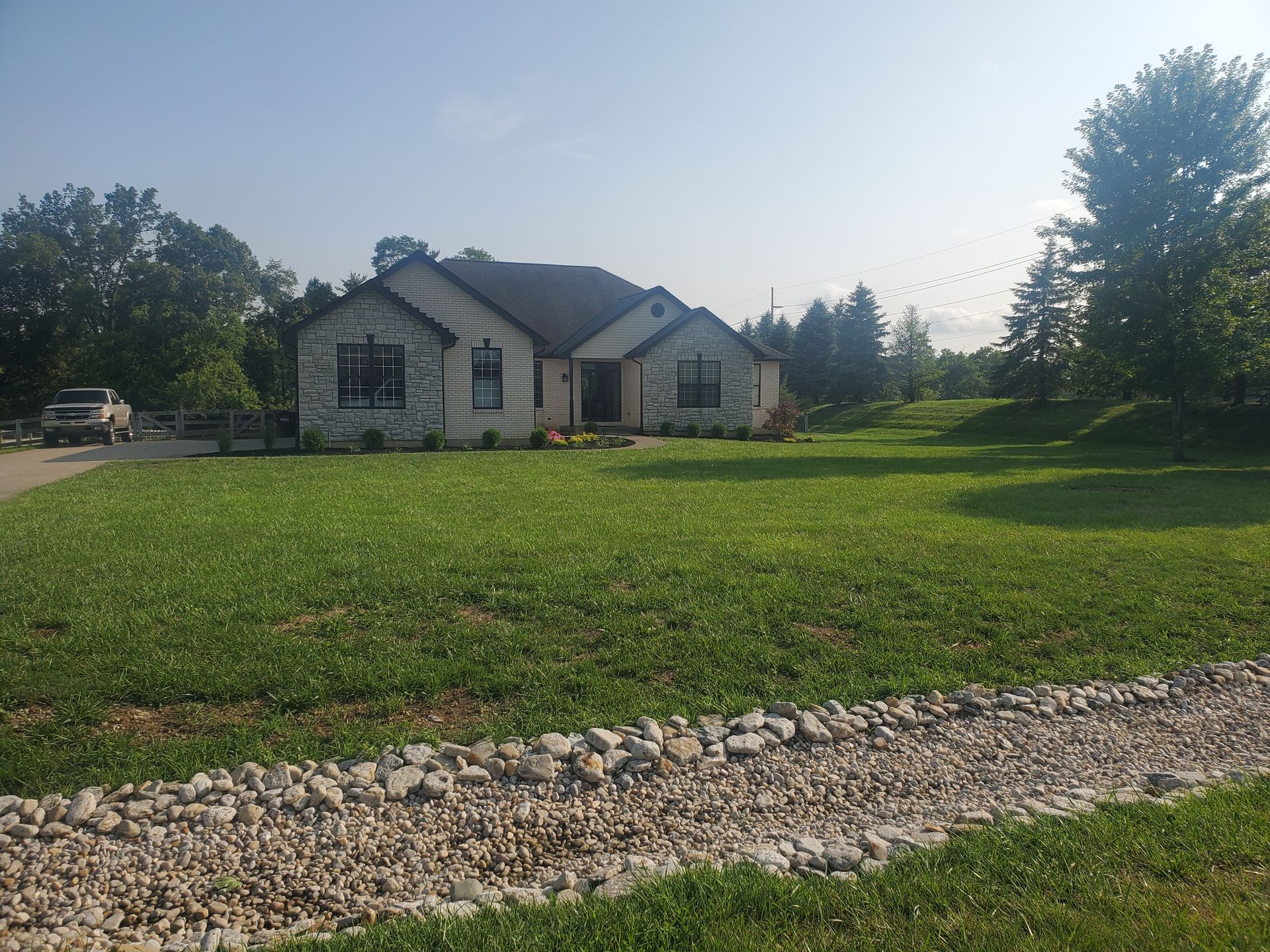 Stone-fronted house with a large green yard, driveway, and rock-lined drainage ditch.