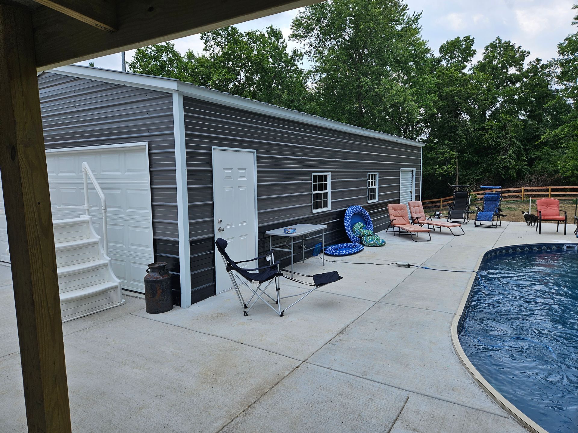 Poolside view of a dark gray garage building with a pool, chairs, and trees in the background.