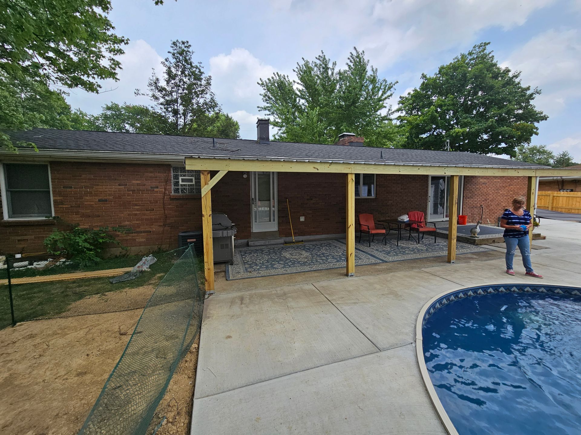 Backyard patio with wooden structure, brick house, concrete and gravel patio. Woman standing. Pool visible.