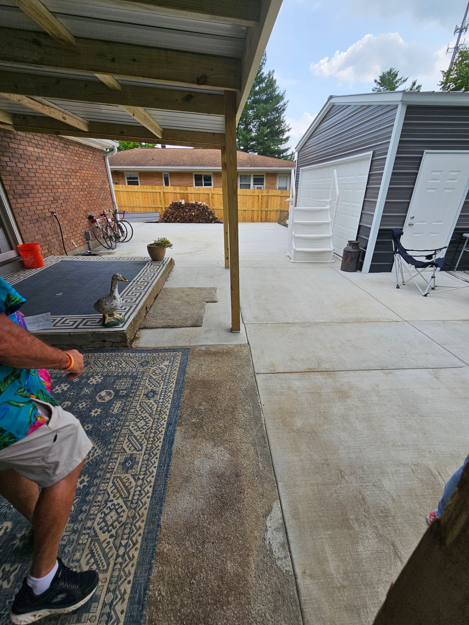 Man holding wood outside, under a deck. Concrete patio and a garage are visible in the background.