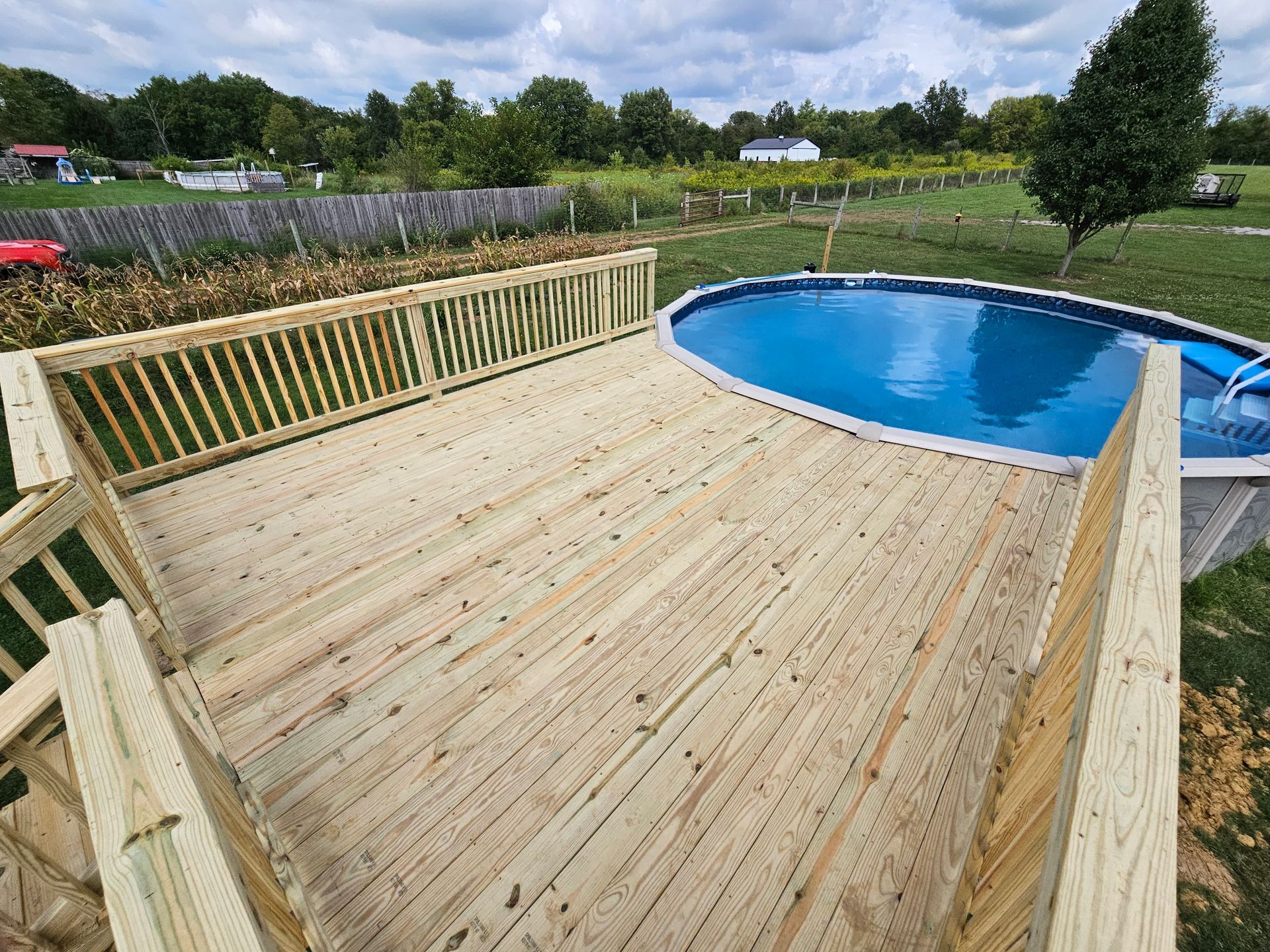 Wooden deck surrounds an oval swimming pool in a grassy yard under a cloudy sky.