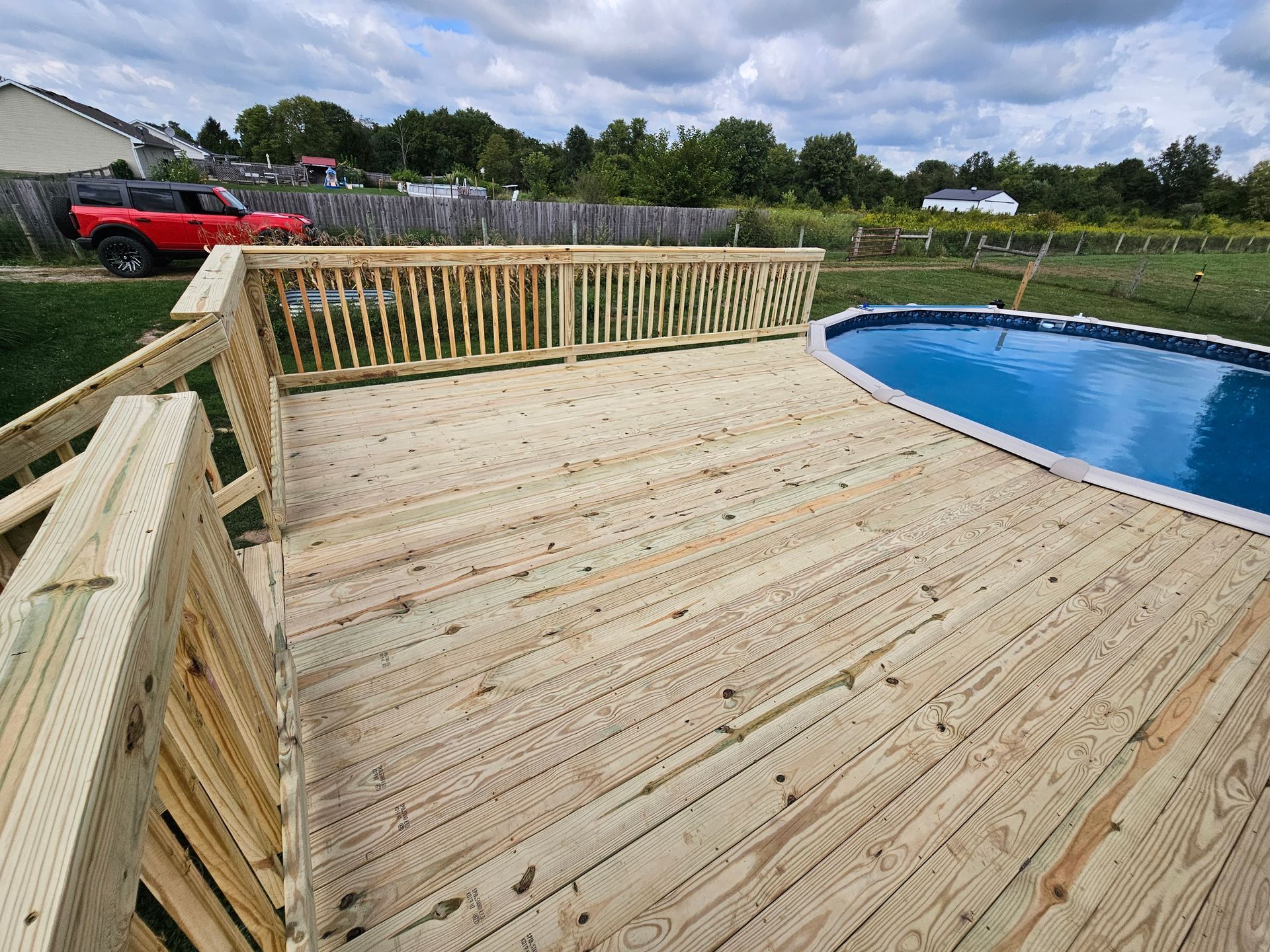 Wooden deck surrounding an oval-shaped pool, overlooking a green yard and cloudy sky.