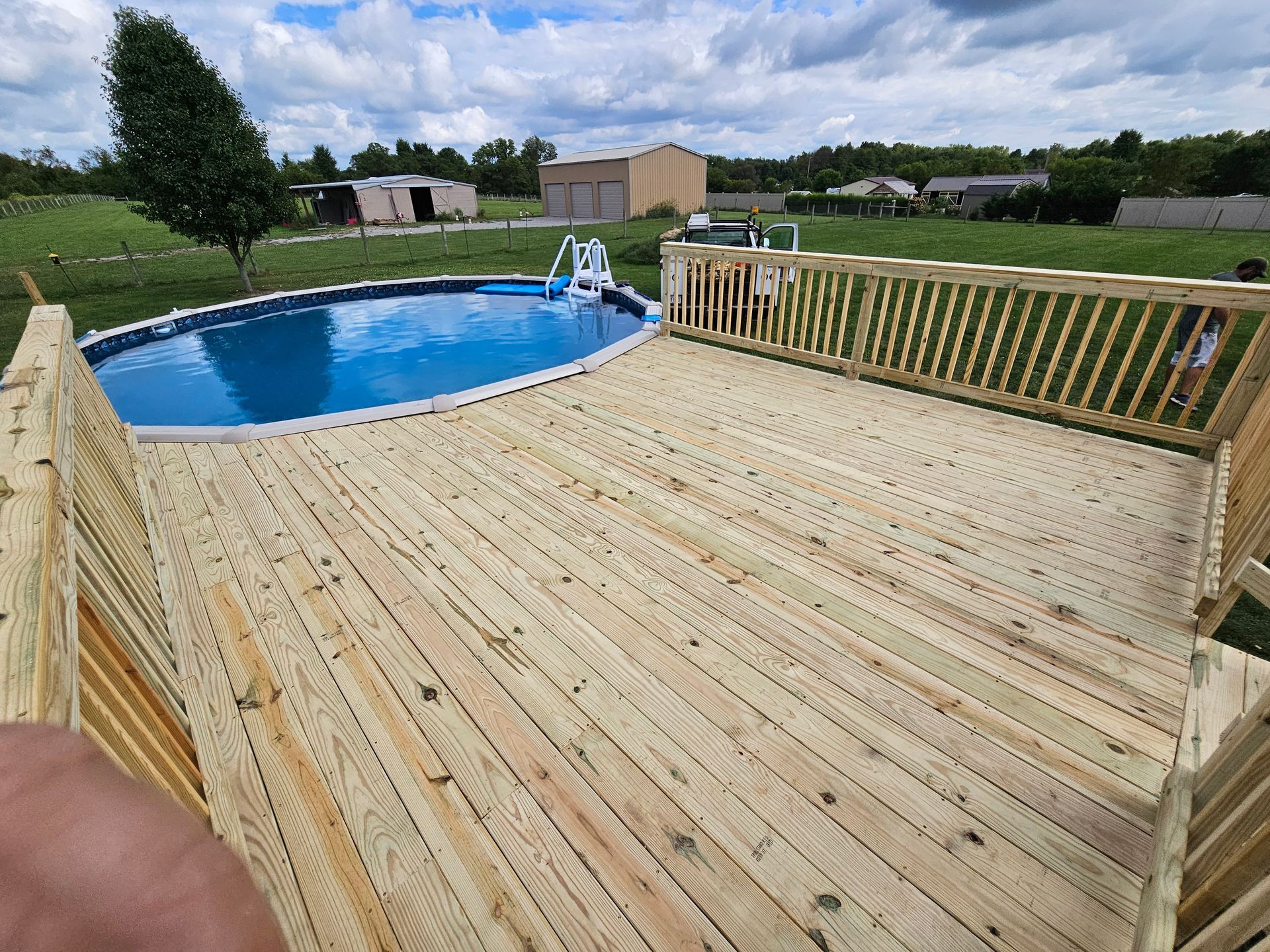 Wooden deck surrounding an above-ground pool. Blue water, clear sky, and rural setting with buildings.