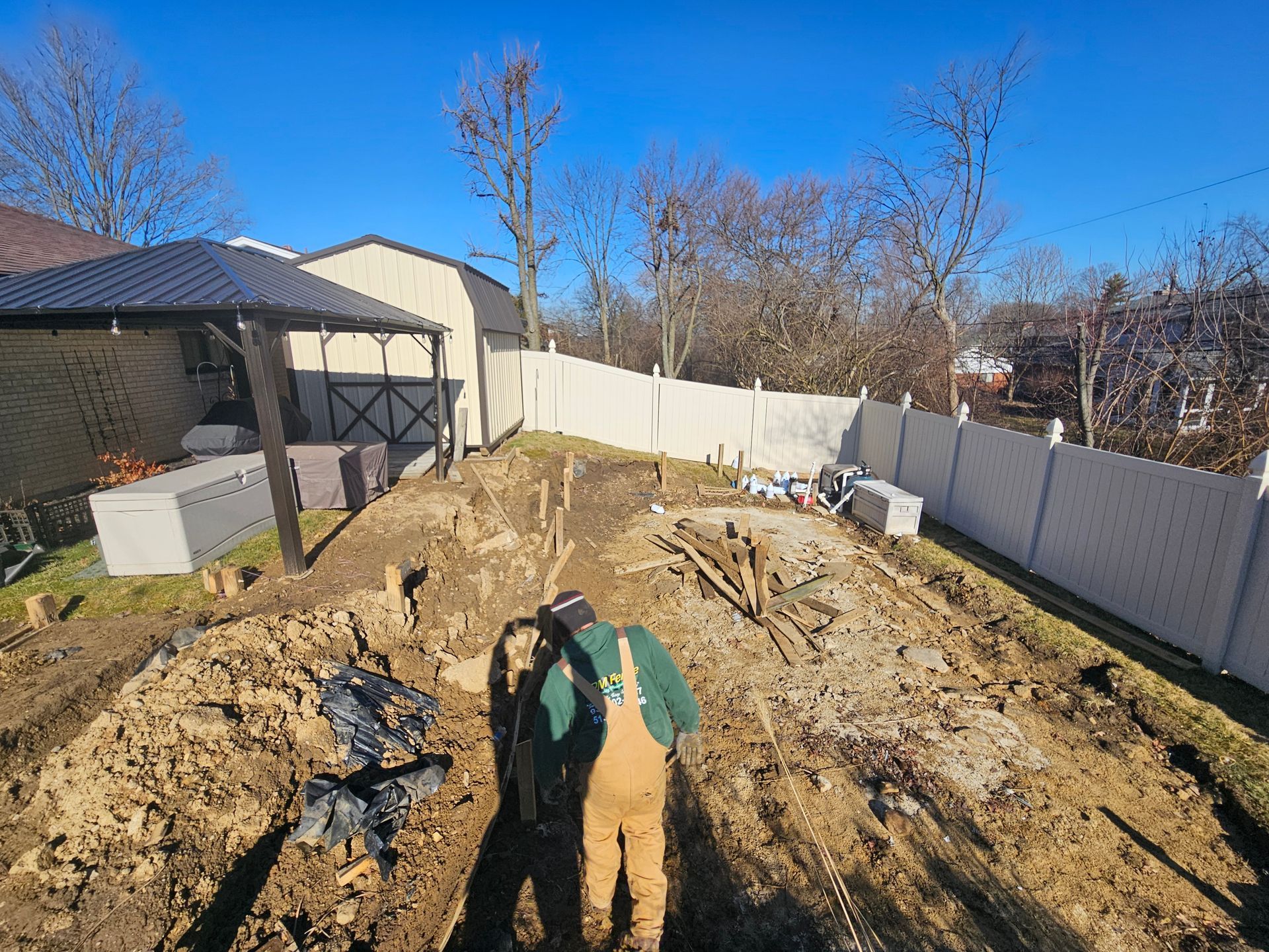 Man in overalls working in a muddy yard, with a fence, shed, and blue sky.