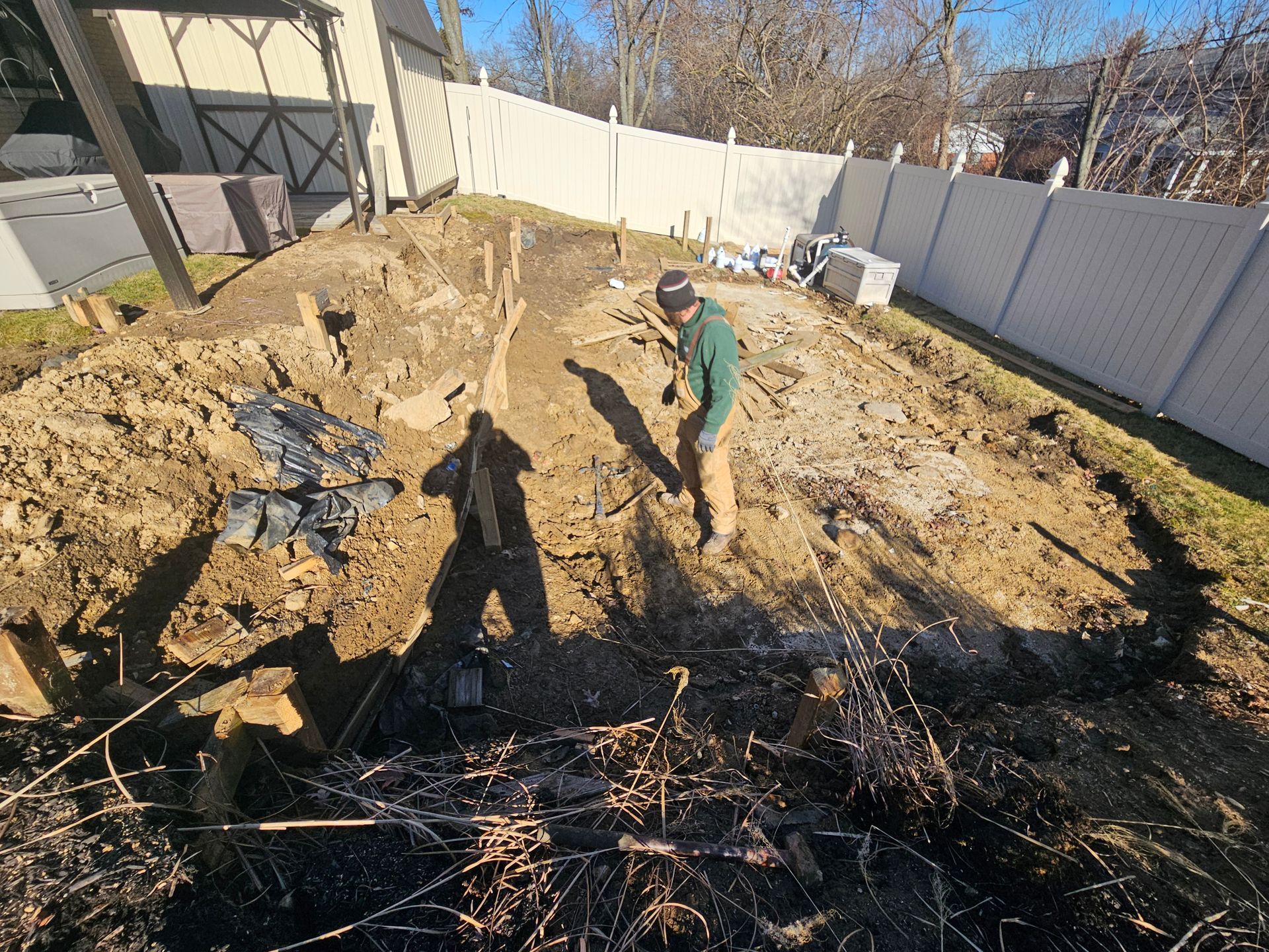 Man digging in a dirt area, preparing for landscaping next to a white fence. Sunny day.