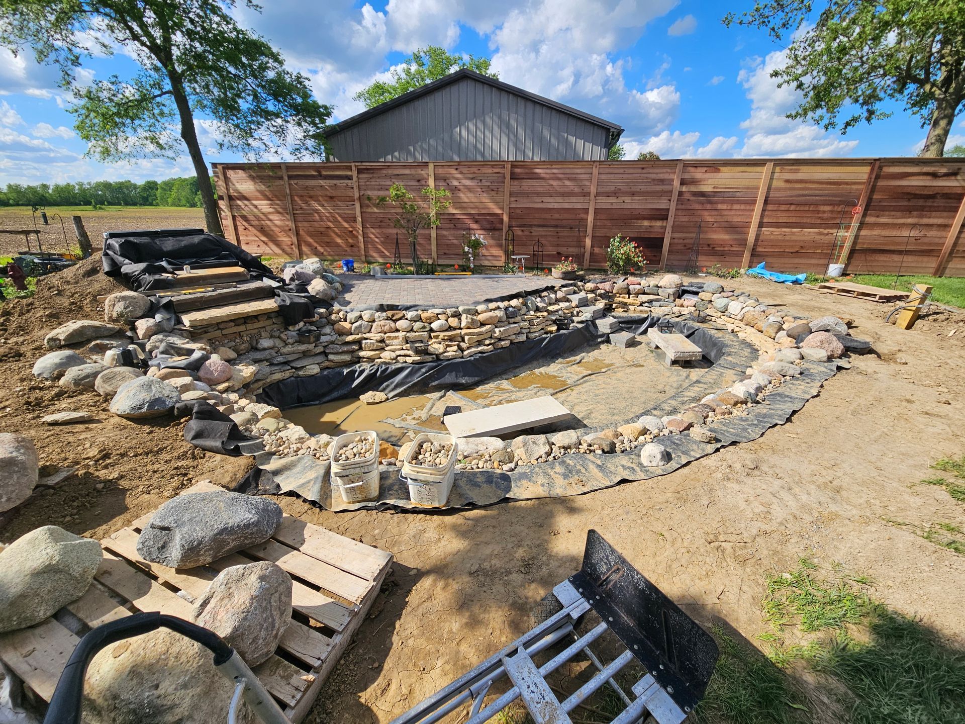 A partially constructed backyard pond with rocks, a black liner, and a wooden fence backdrop under a blue sky.