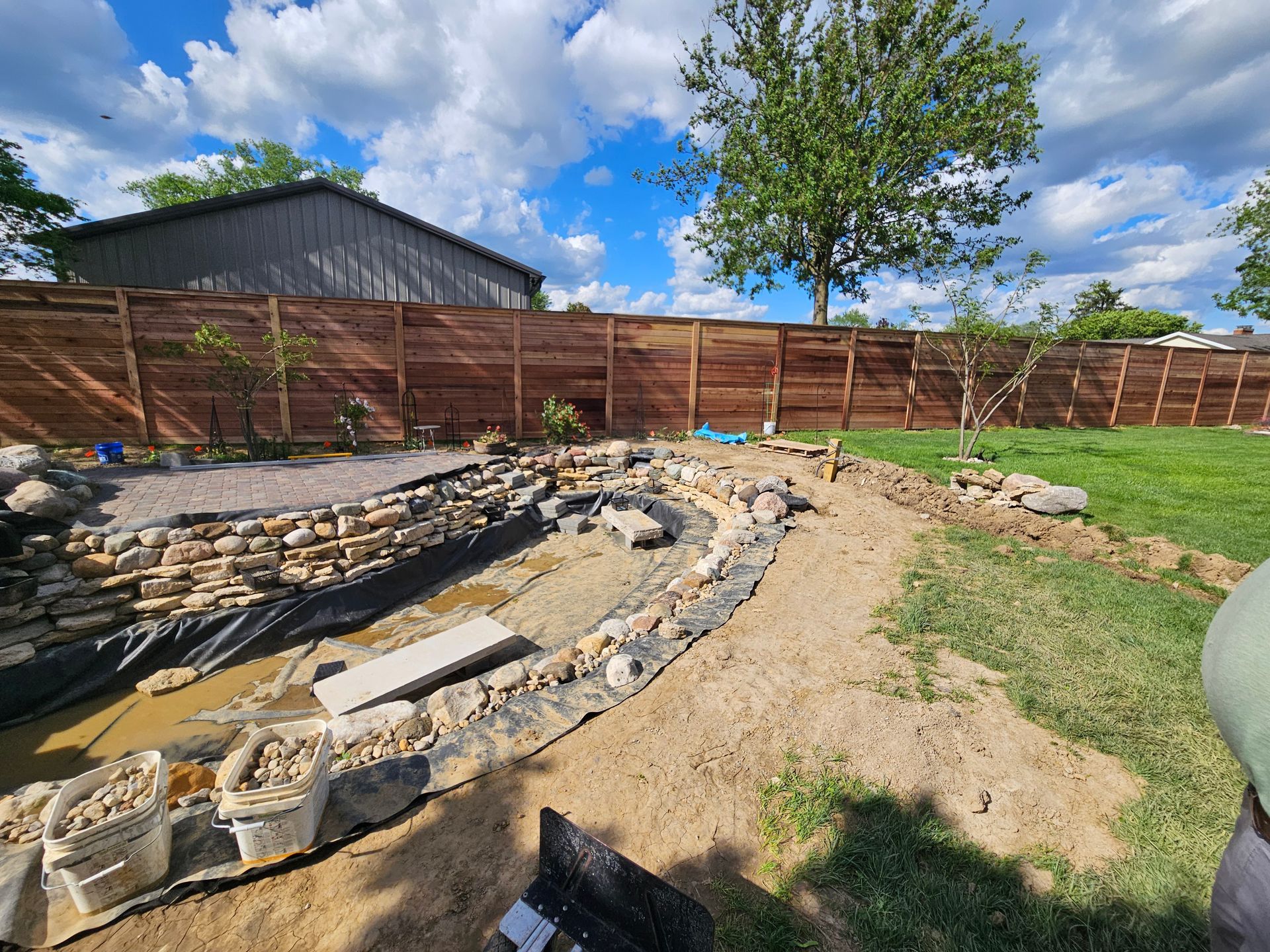 Backyard with a pond under construction. Brown fence, blue sky with clouds, grass and dirt.