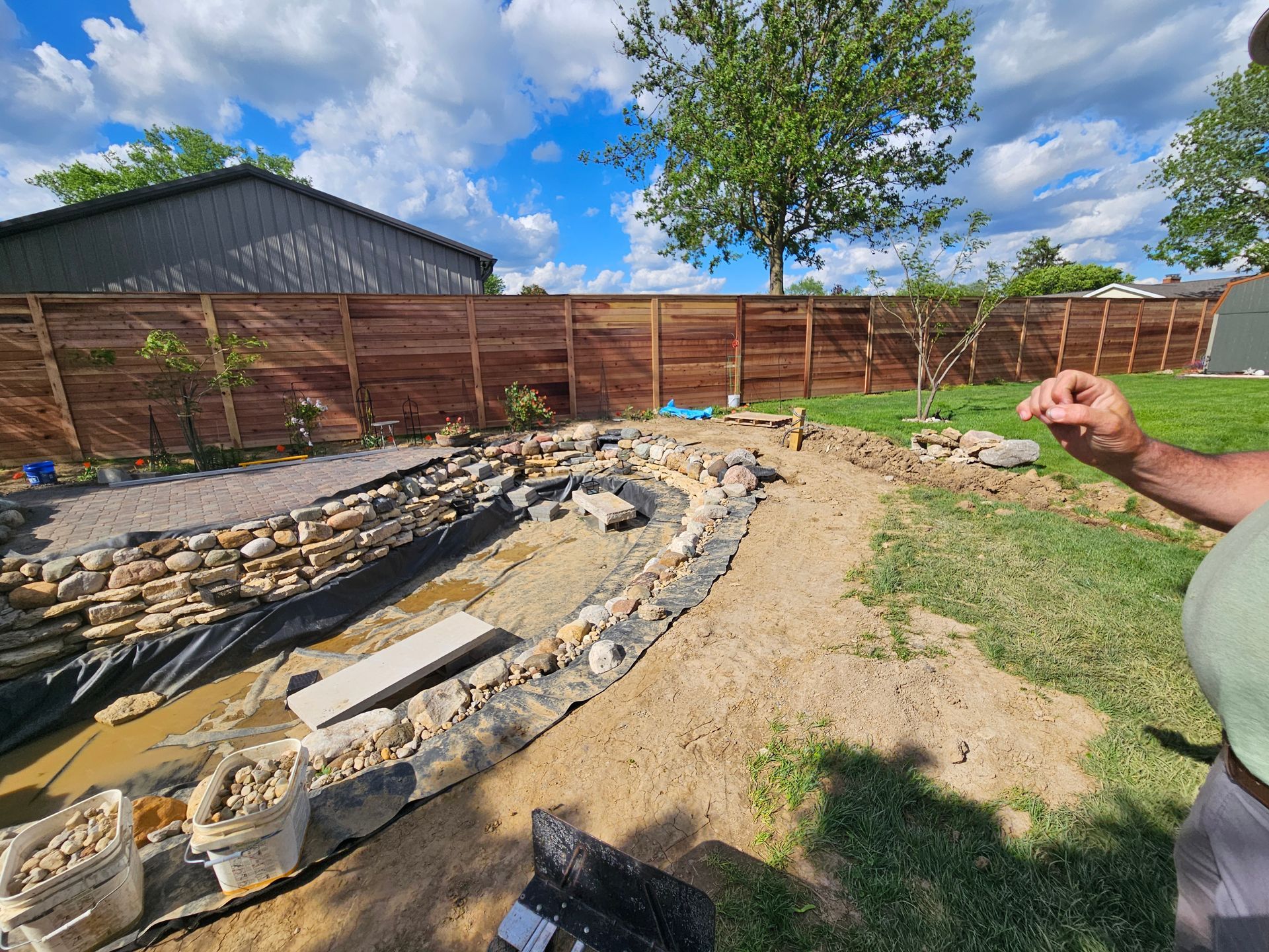 A backyard pond under construction with rocks, dirt, and a wooden fence in the background on a sunny day.