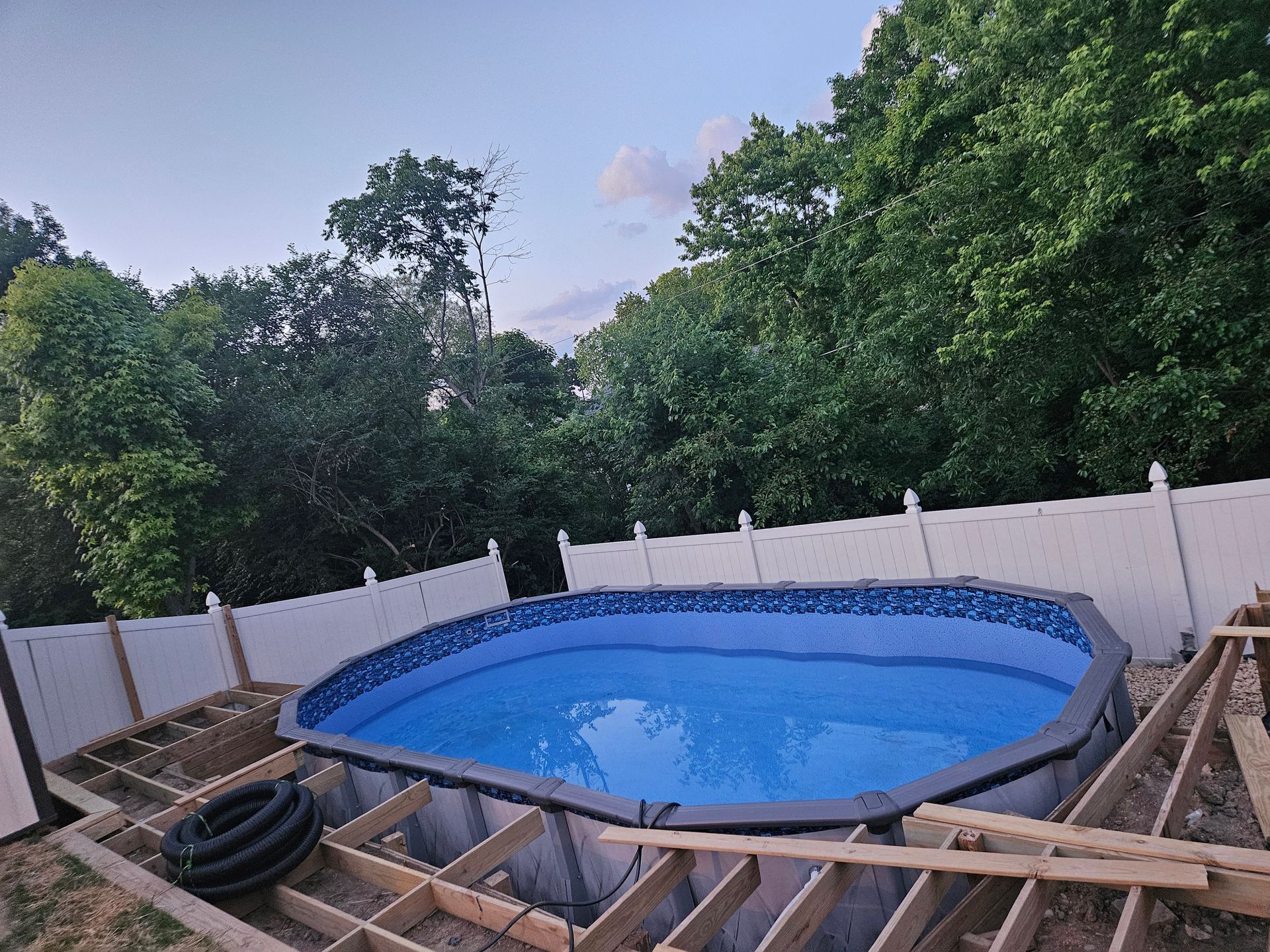 Above-ground pool surrounded by unfinished wooden deck and white fence; trees in background.