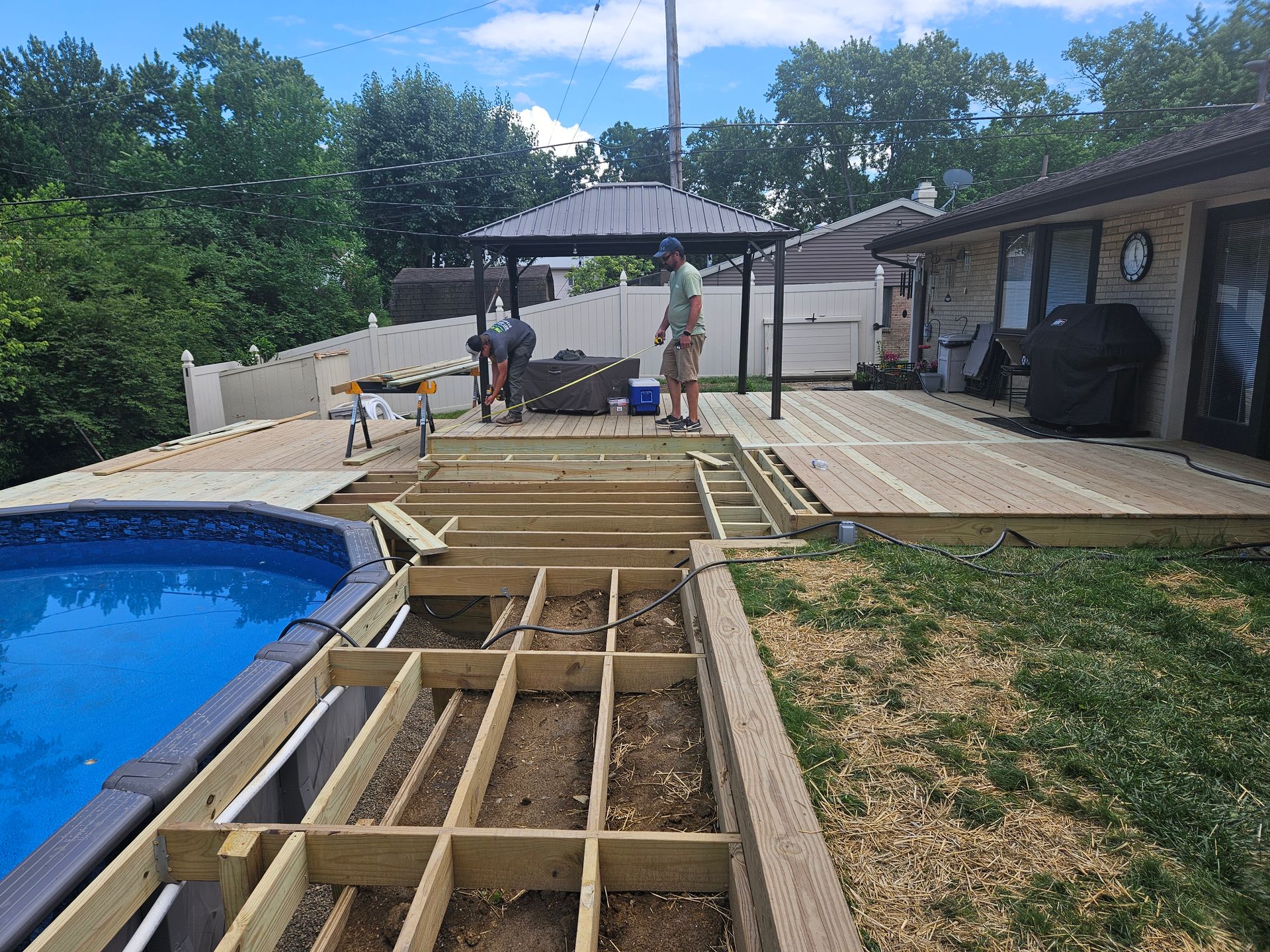 Deck construction near a pool; two workers, gazebo, green grass, blue pool.
