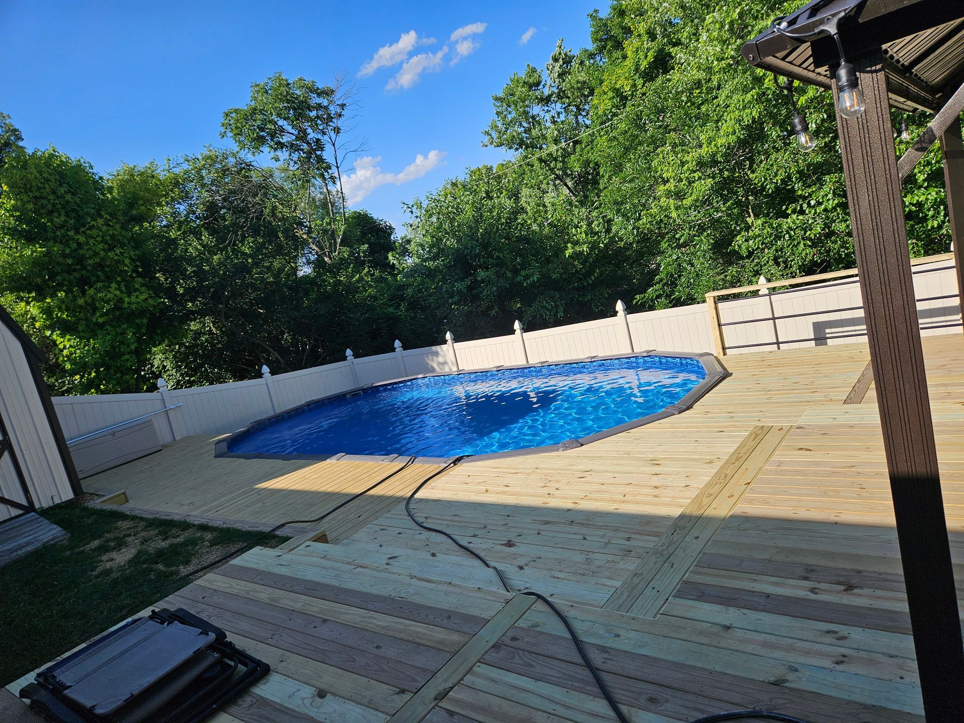 Wooden deck with an oval pool covered in blue, surrounded by a white fence and trees.