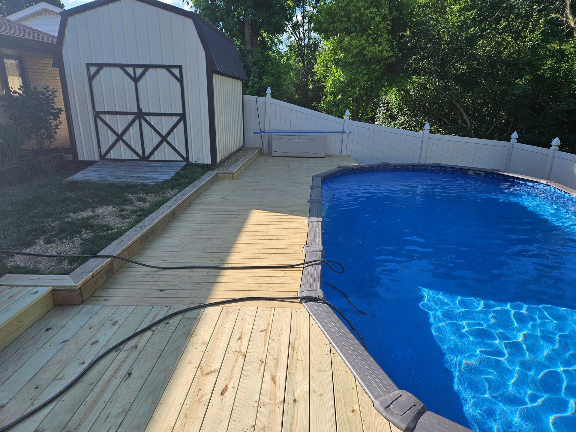 Wooden deck next to a blue swimming pool, a shed, and white fence.