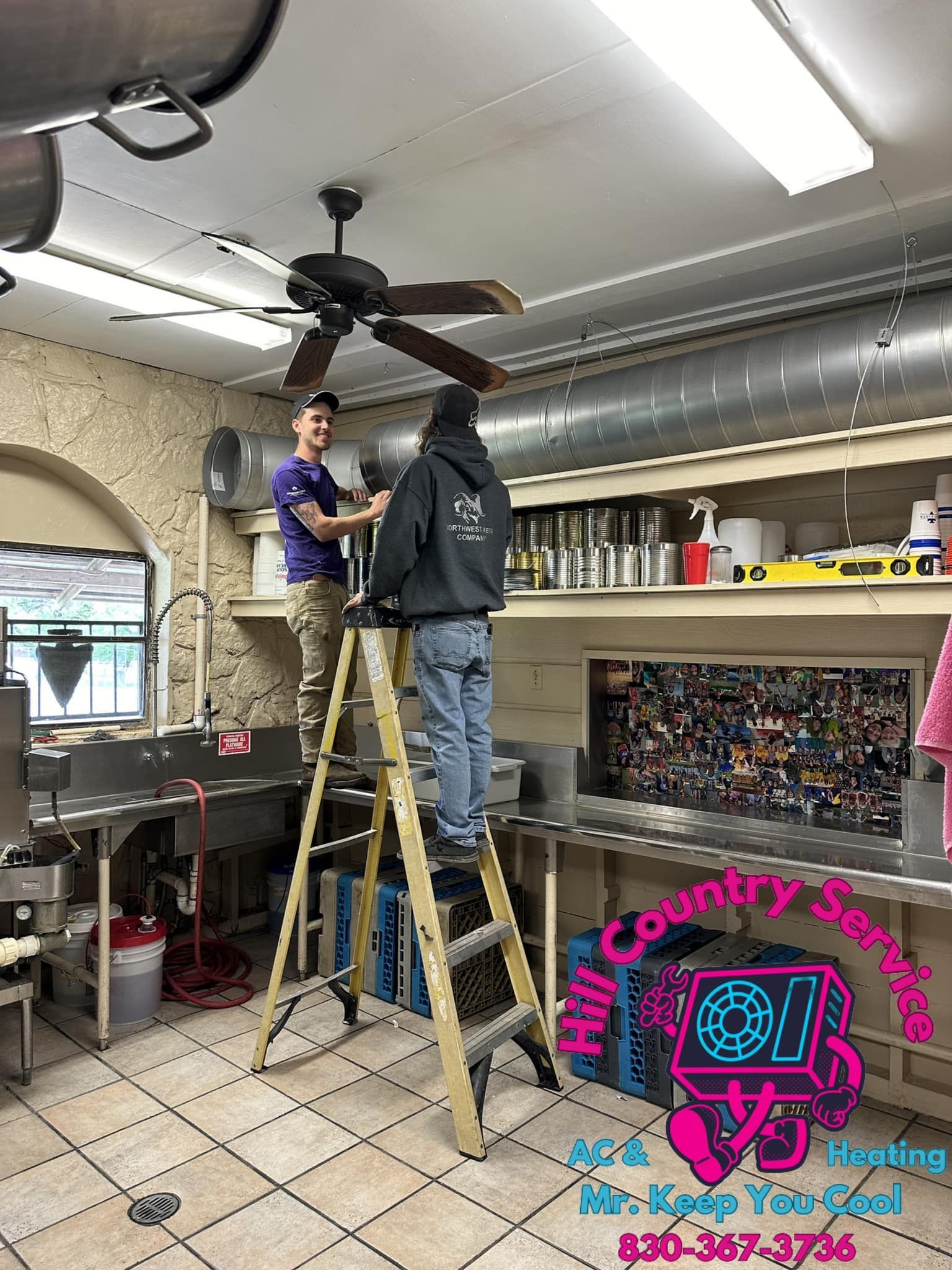 Two men are working on a ceiling fan in a kitchen.