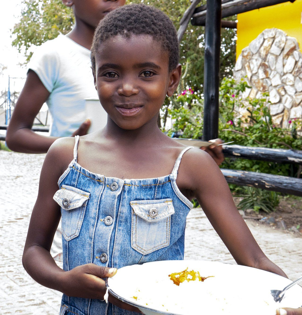 A young girl in a denim dress is holding a plate of food
