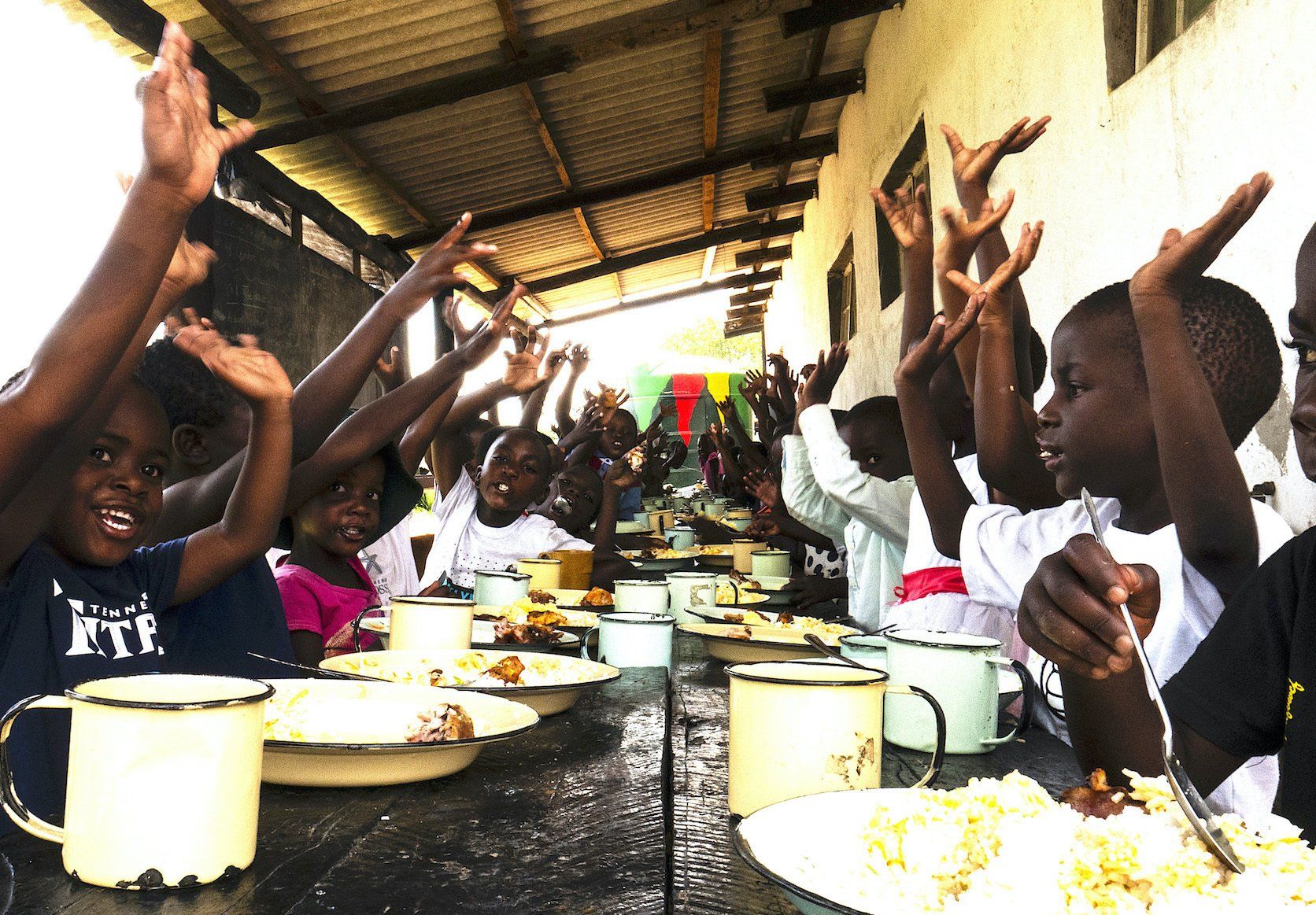 A group of children are sitting at a table with their hands in the air.