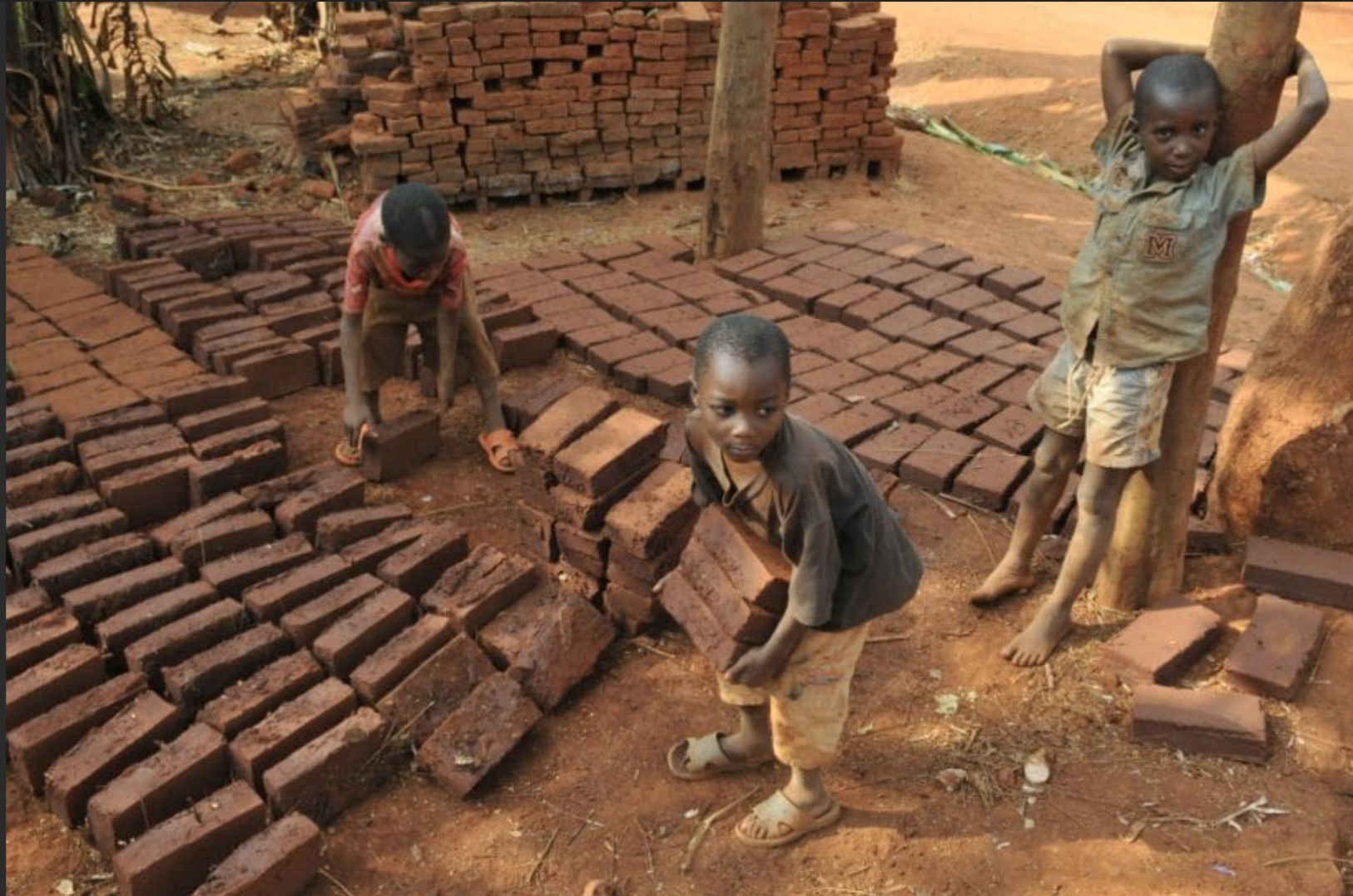 Children stacking bricks in a dusty, outdoor area. One child carries a brick, two others work nearby.