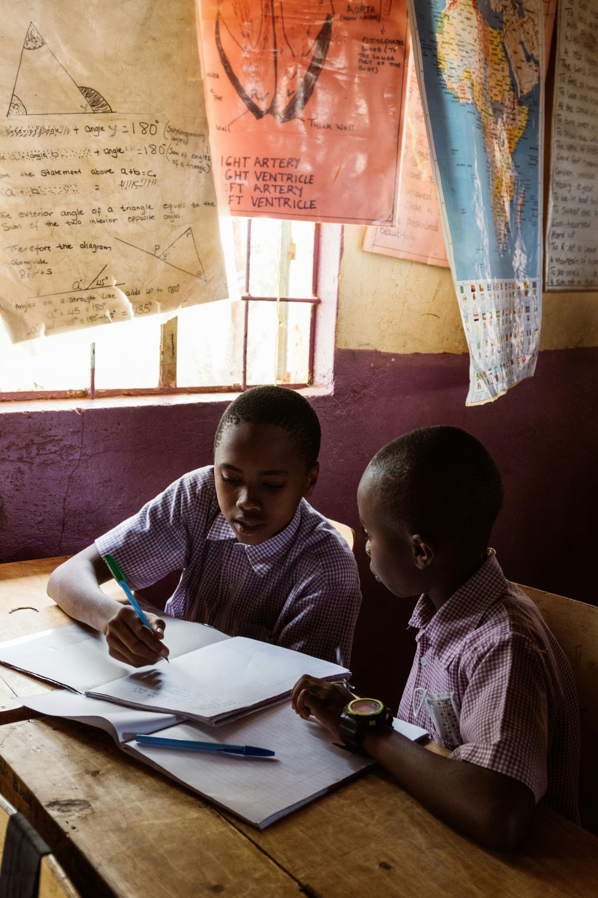 Two students studying together at a desk in a classroom, writing in notebooks.