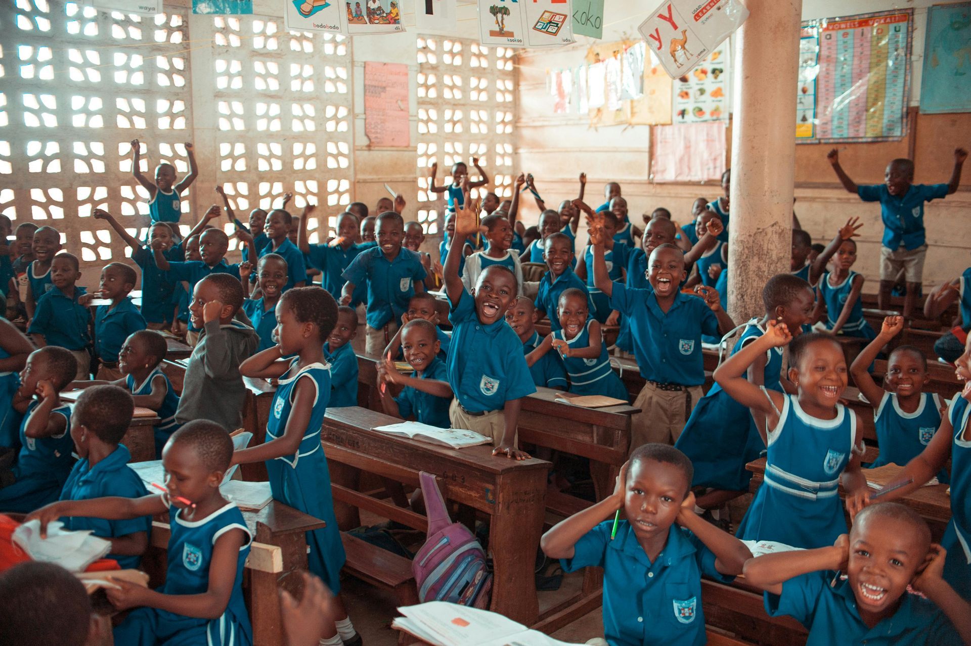 Children in blue uniforms in a classroom with raised arms and happy expressions. Wooden desks, posters.