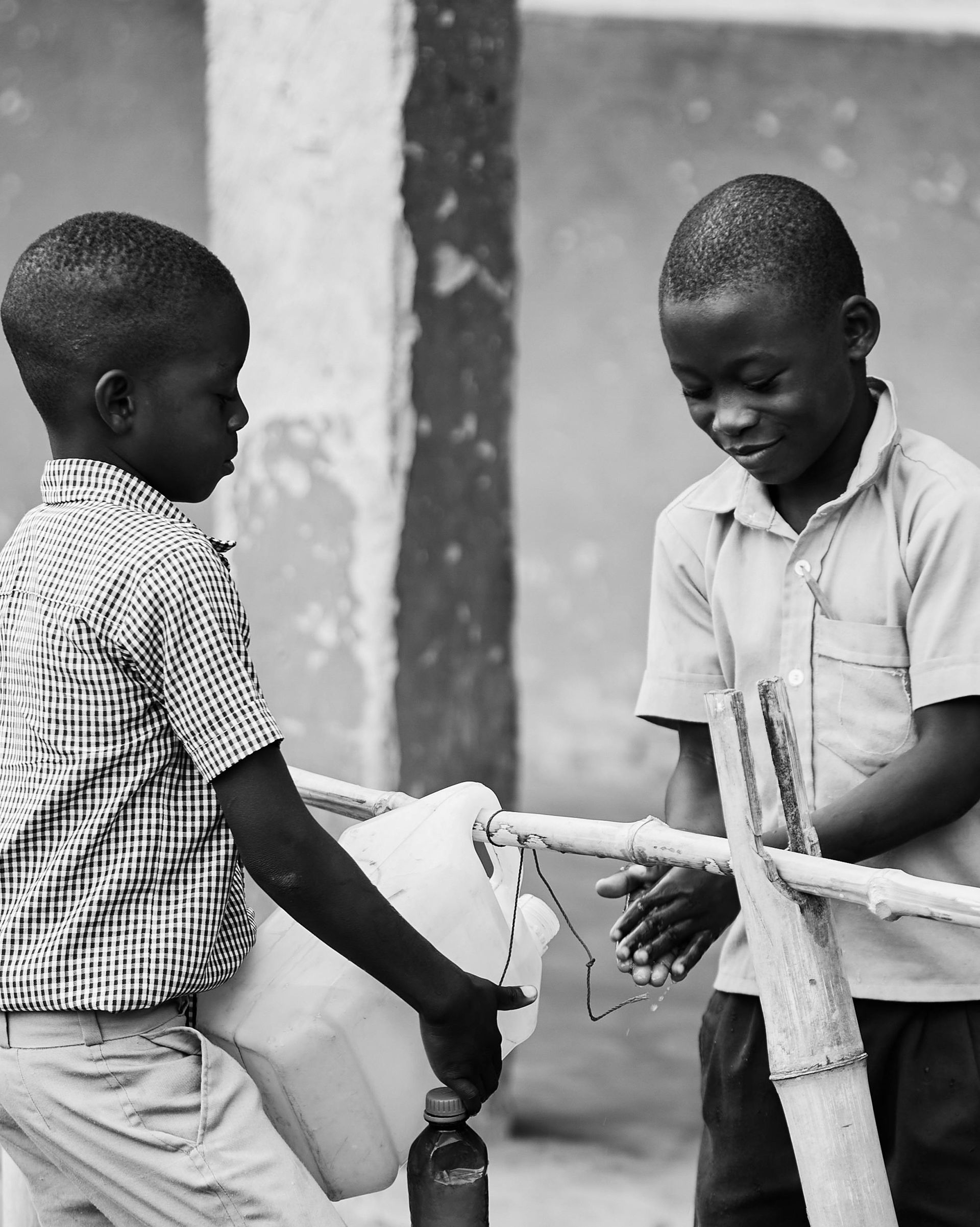 Two boys washing hands at a makeshift handwashing station outdoors, one pouring water.