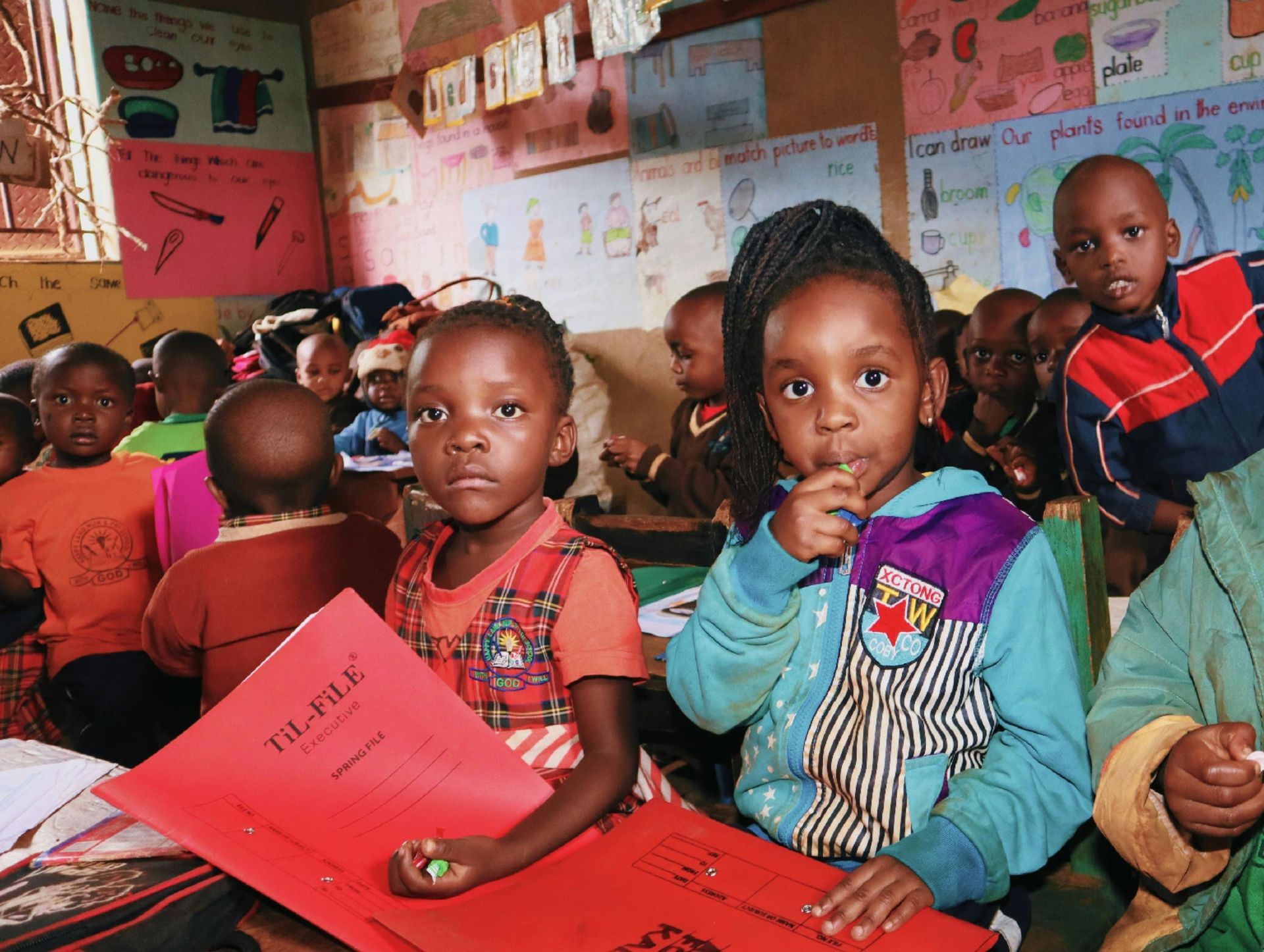 Children in a colorful classroom, two girls holding red folders looking toward the viewer.