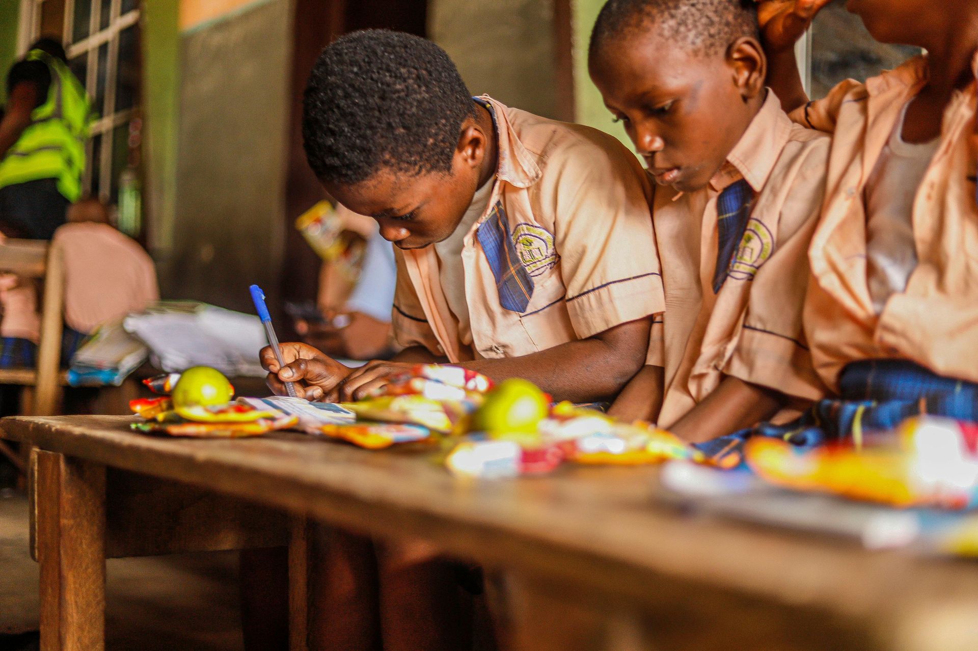 Students in uniform write at a desk with lunch, in a classroom.