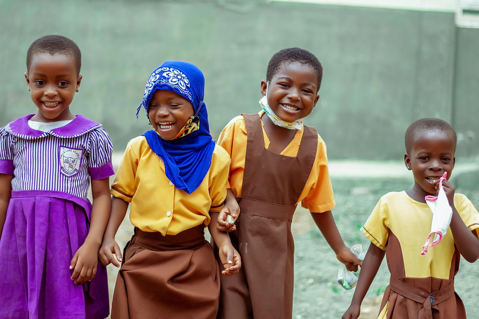 Four smiling school children wearing uniforms. They hold hands, outdoors.