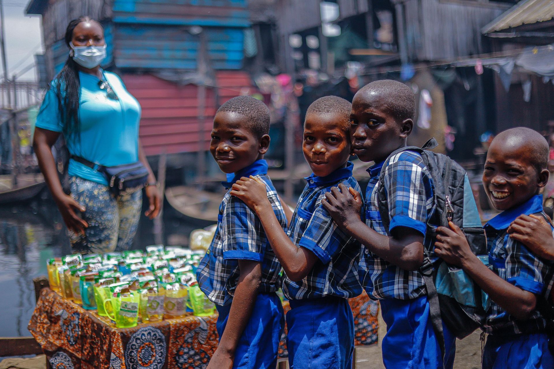 Woman in mask with school children in uniform near a table of products; outdoor, slum setting.