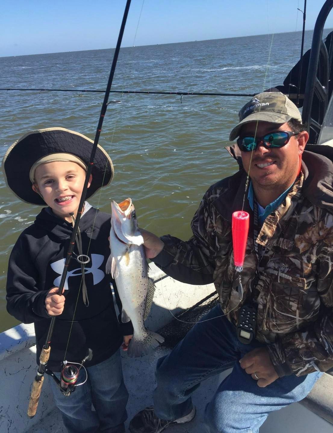 A man and a boy are fishing on a boat and the boy is wearing a pirate hat.