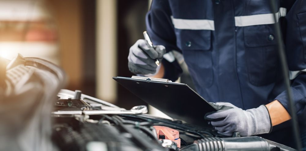 A mechanic is working on a car in a garage while holding a clipboard and a pen.