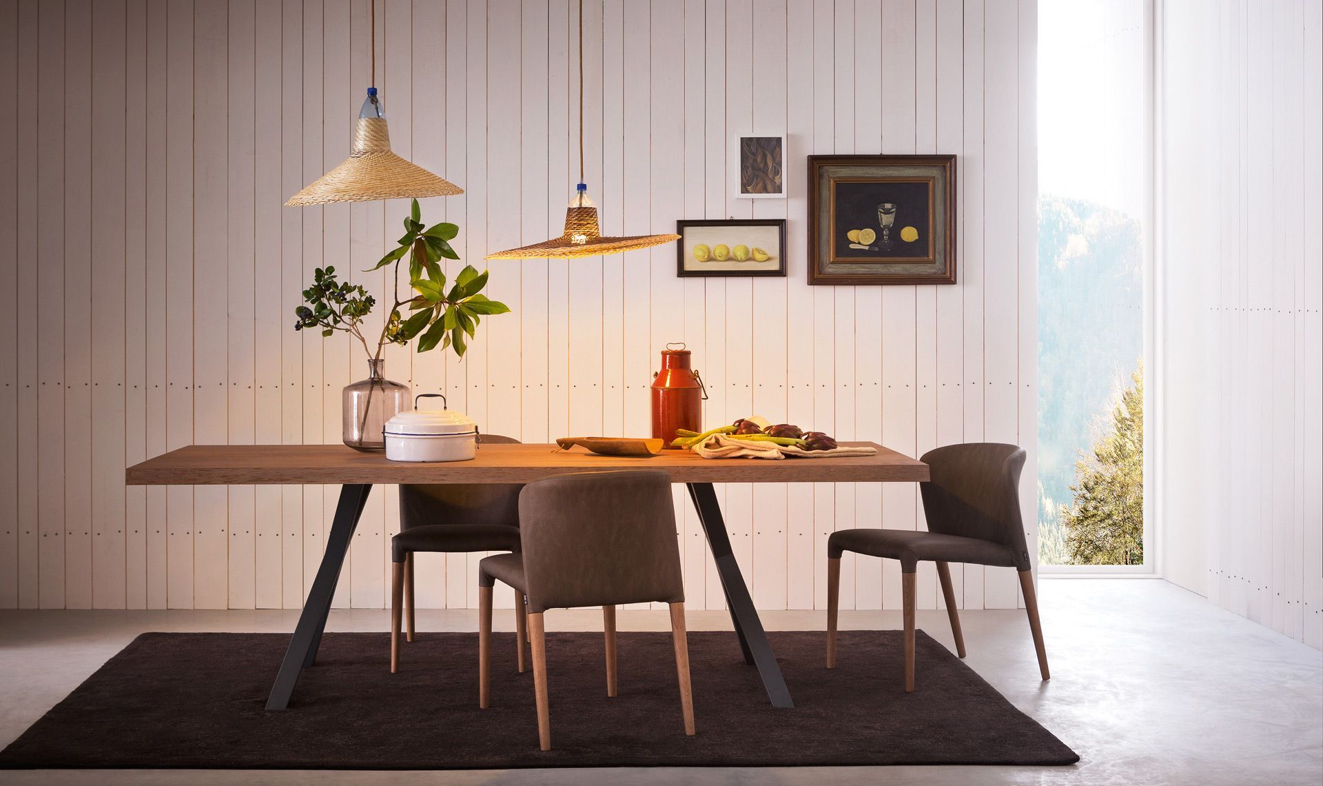 Sala da pranzo con tavolo in legno, sedie scure e lampade intrecciate. Opere d'arte appese su una parete rivestita di pannelli bianchi.