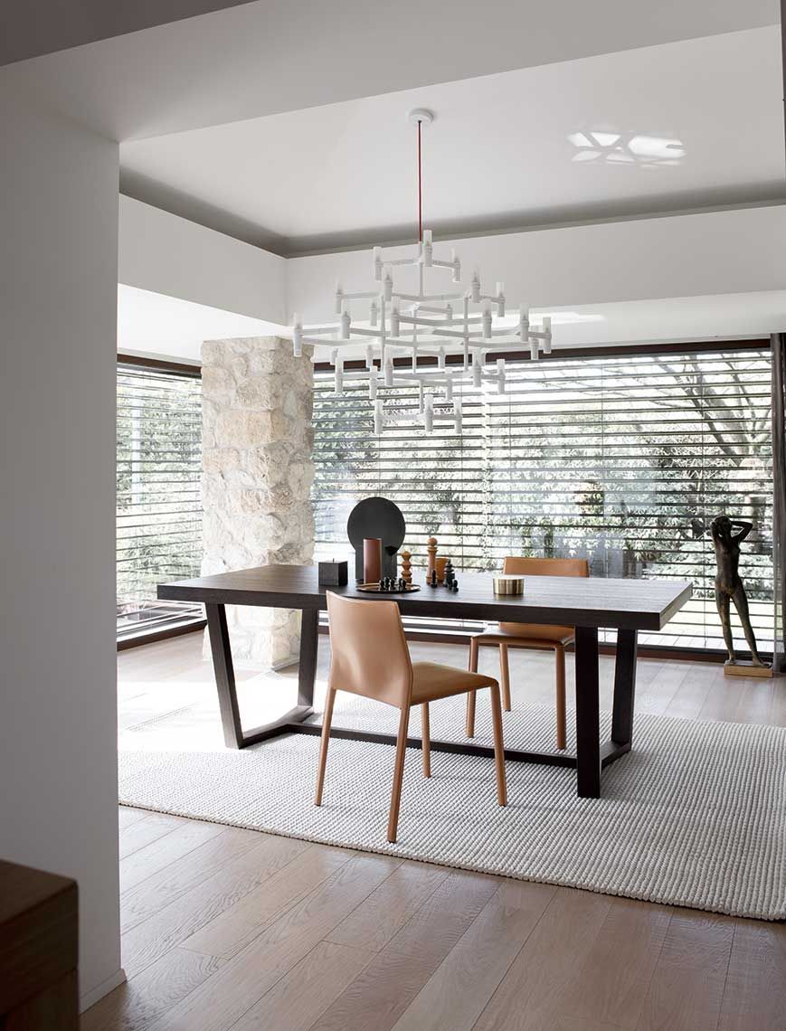 Sala da pranzo con grande tavolo in legno scuro, sedie chiare, tappeto a fantasia e lampadario geometrico.