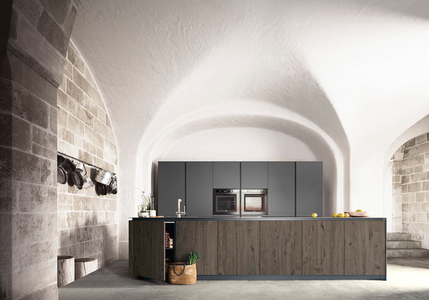 Cucina moderna in una stanza con pareti in pietra e arco, isola in legno, mobili grigi ed elettrodomestici da incasso.