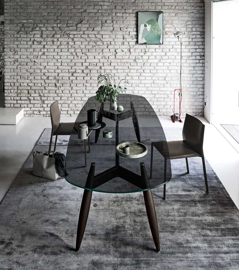 Tavolo da pranzo ovale in vetro con gambe in legno scuro, due sedie marroni e tappeto contro una parete di mattoni bianchi.
