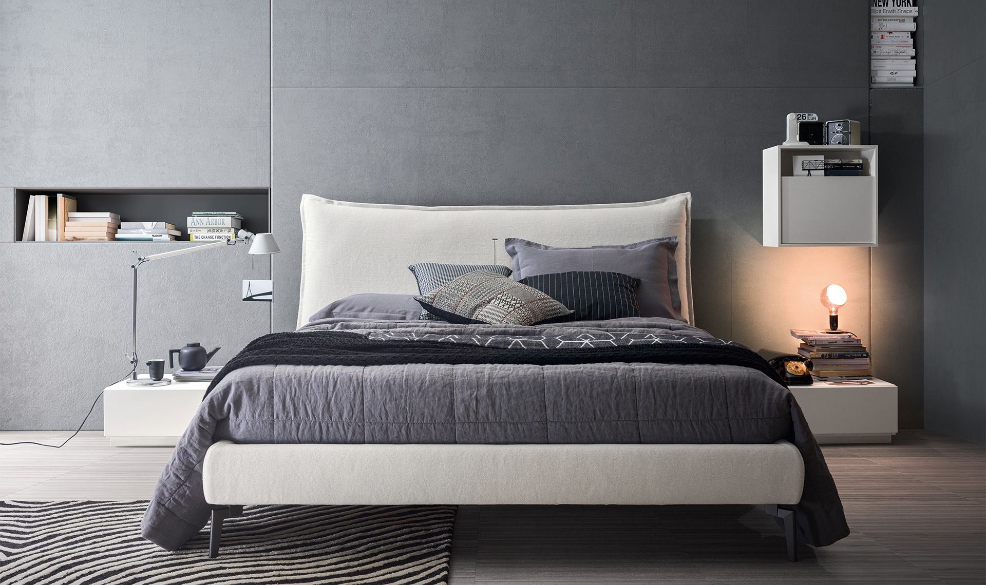 Camera da letto moderna con letto, piumone grigio e testiera bianca, accanto a comodini sospesi.