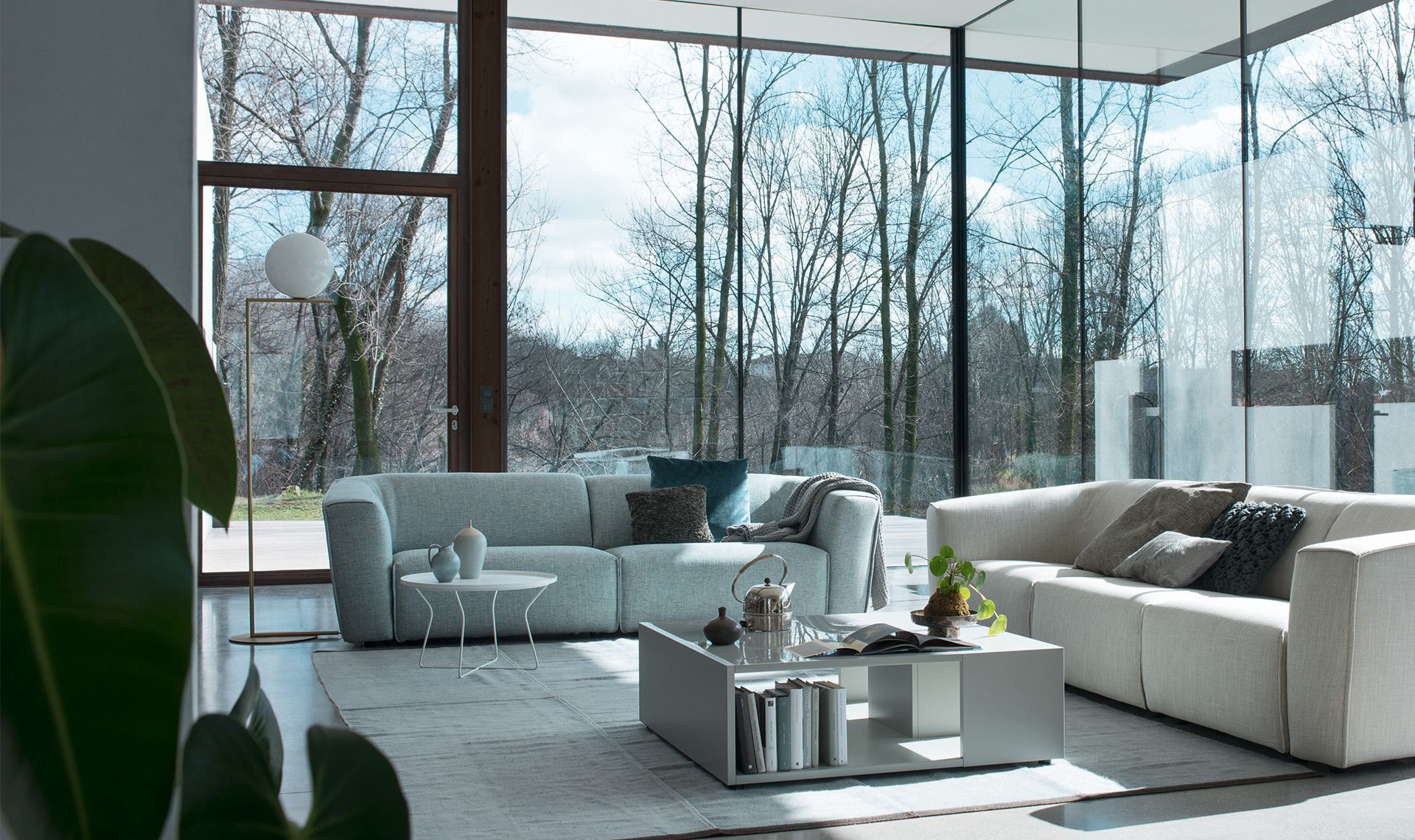 Soggiorno moderno con divani dai colori chiari, ampie finestre con vista sugli alberi e un tavolino da caffè bianco.