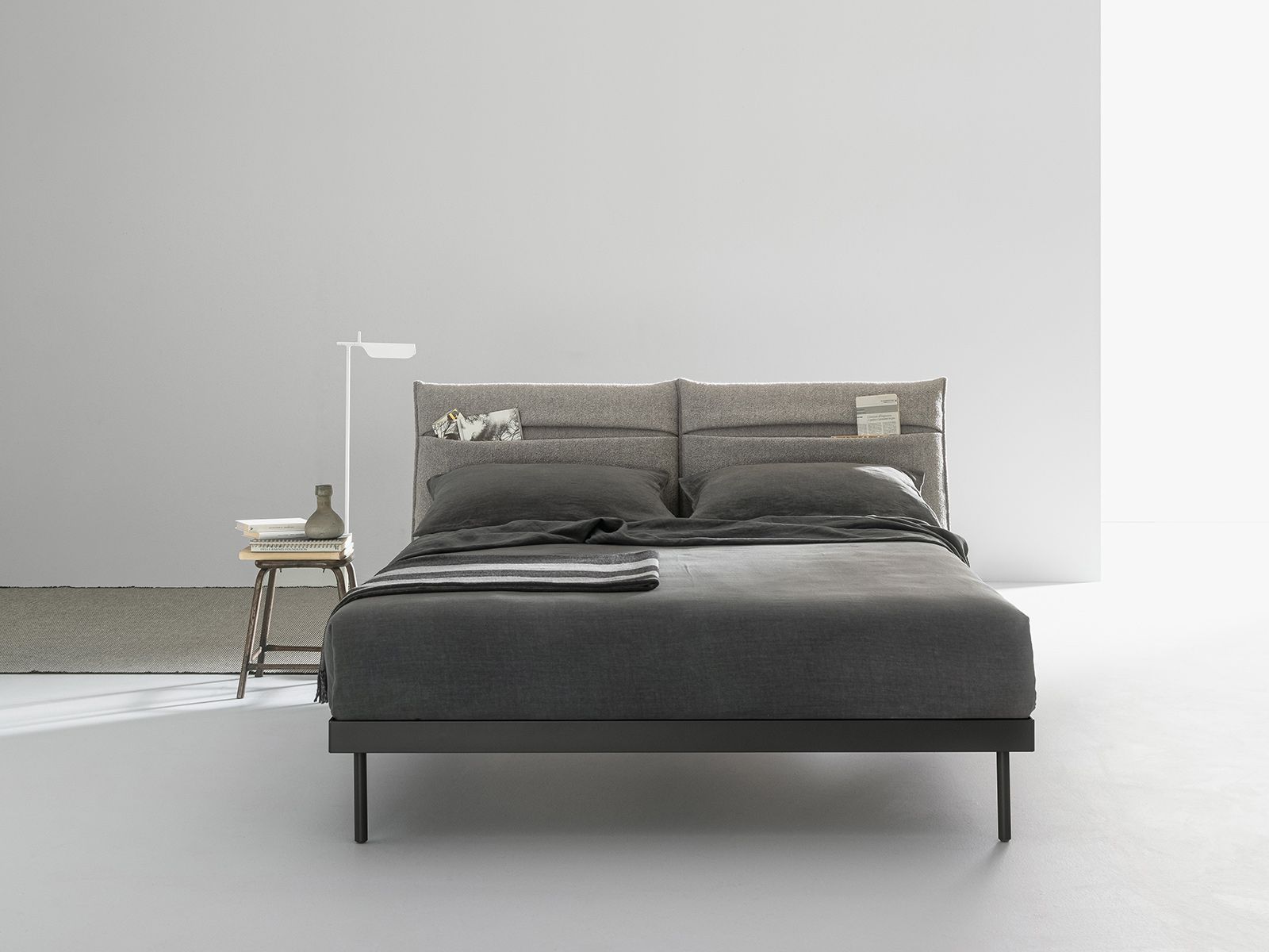 Letto grigio con testiera testurizzata, coperta, cuscini, tavolino con lampada, sfondo neutro.