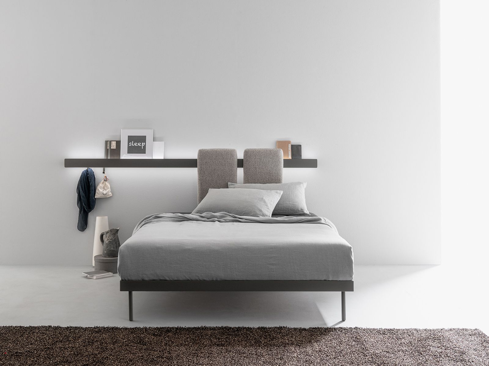 Letto moderno con biancheria grigia, testiera con mensola e oggetti, struttura scura, su un tappeto.