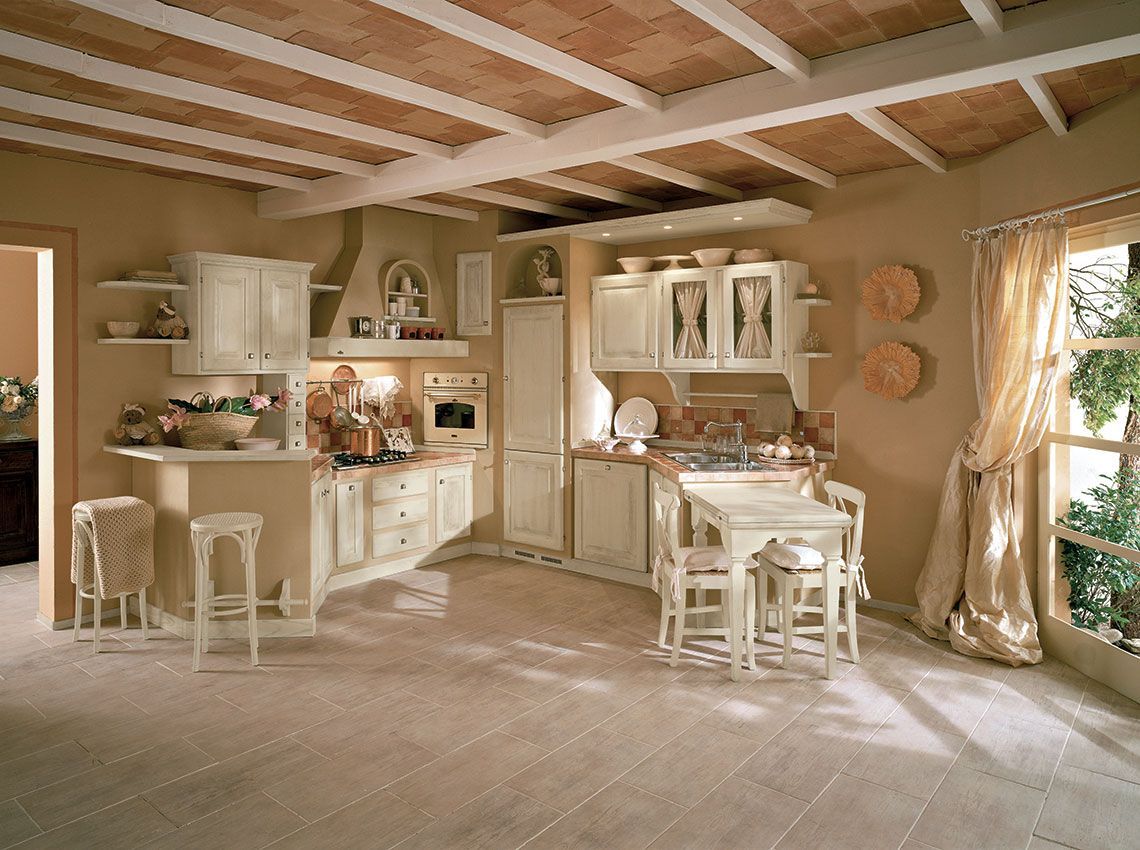 Cucina color crema con mobili, un tavolo e due sgabelli. Soffitto con travi a vista e finestra con tenda.