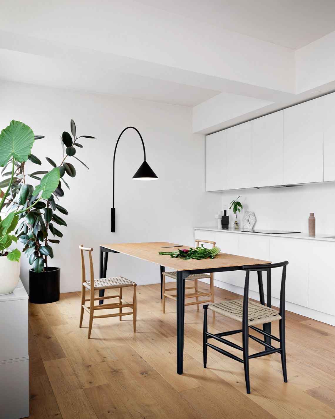 Zona pranzo con tavolo in legno, sedie nere e lampada moderna contro la parete bianca; mobili da cucina sullo sfondo.