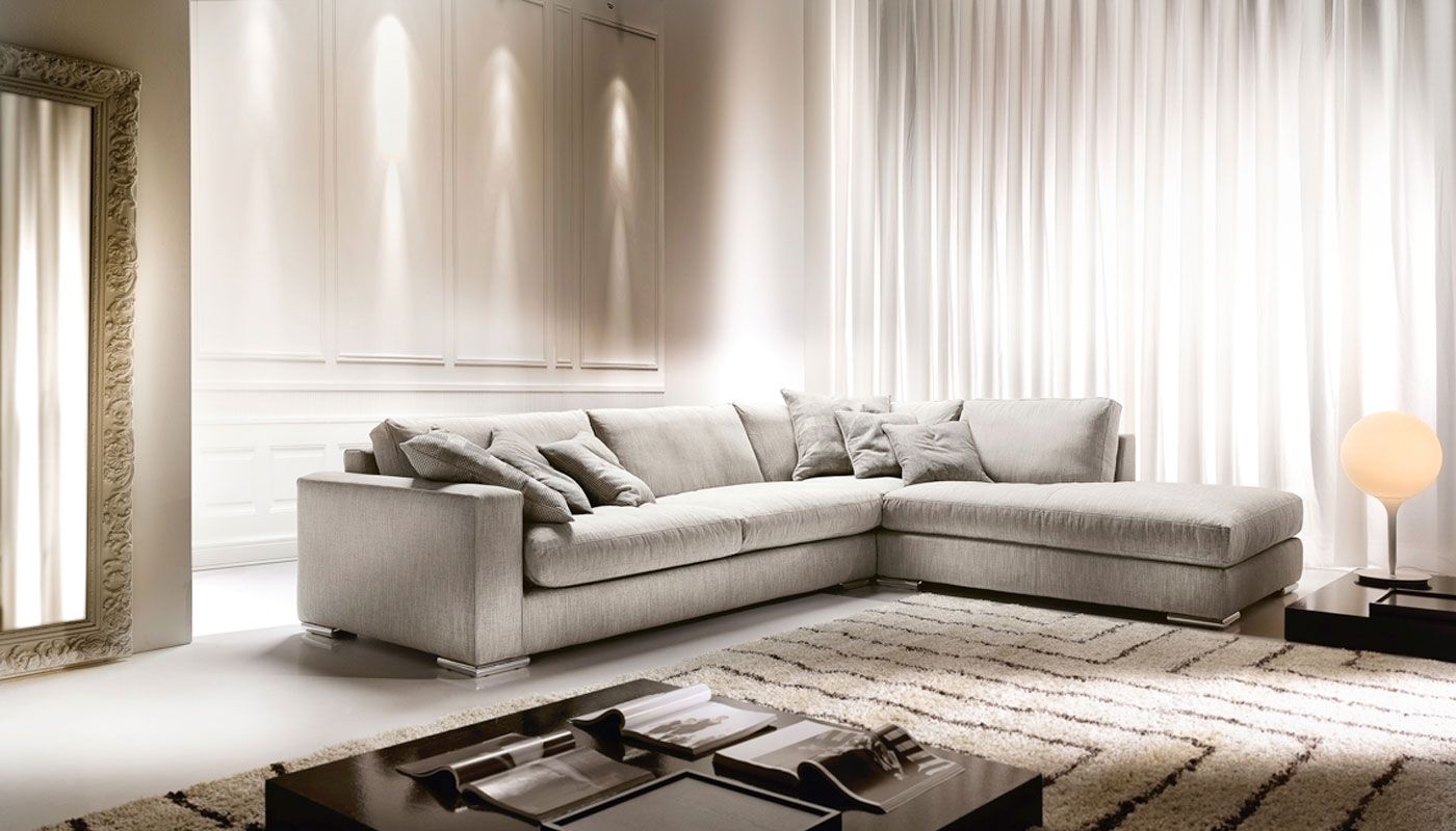 Divano beige a L in un soggiorno moderno, con tende bianche, un tappeto e uno specchio.