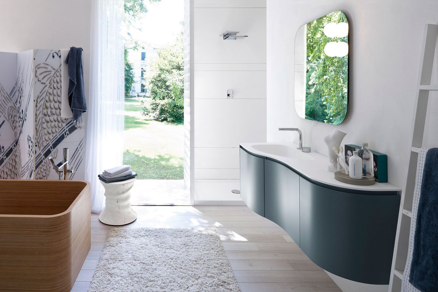 Bagno moderno con vasca in legno, mobile lavabo curvo blu, grande specchio e finestra aperta sul giardino.