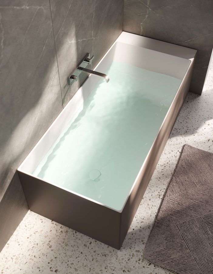 Vasca da bagno rettangolare piena d'acqua; rubinetto su una parete piastrellata grigia. Tappetino da bagno beige sul pavimento.