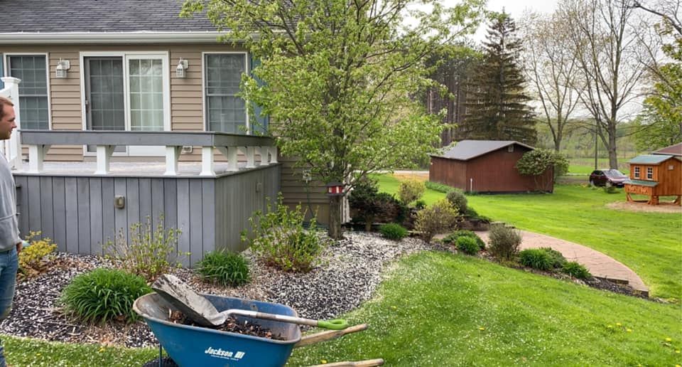 A man is standing in front of a house with a wheelbarrow full of rocks.