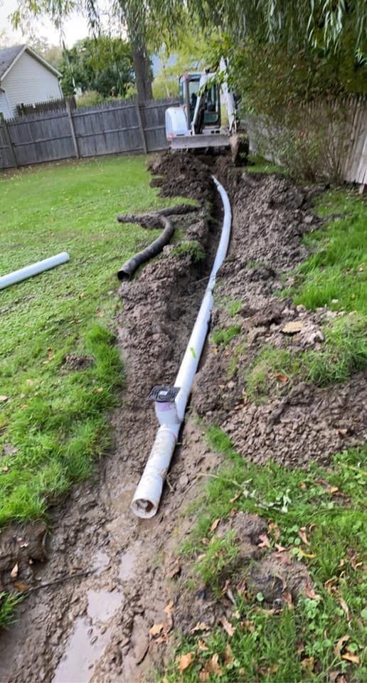 A trench with white drainage pipes and a small backhoe in a yard.