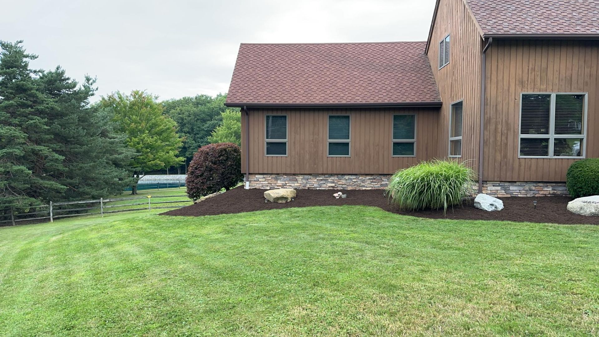 A wooden house with a brown shingled roof, windows, and manicured landscaping, set on a green lawn by a treeline.