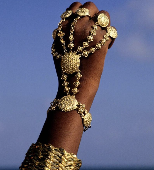 Gold hand jewelry against a blue sky, including a hand chain, bracelet, and rings.