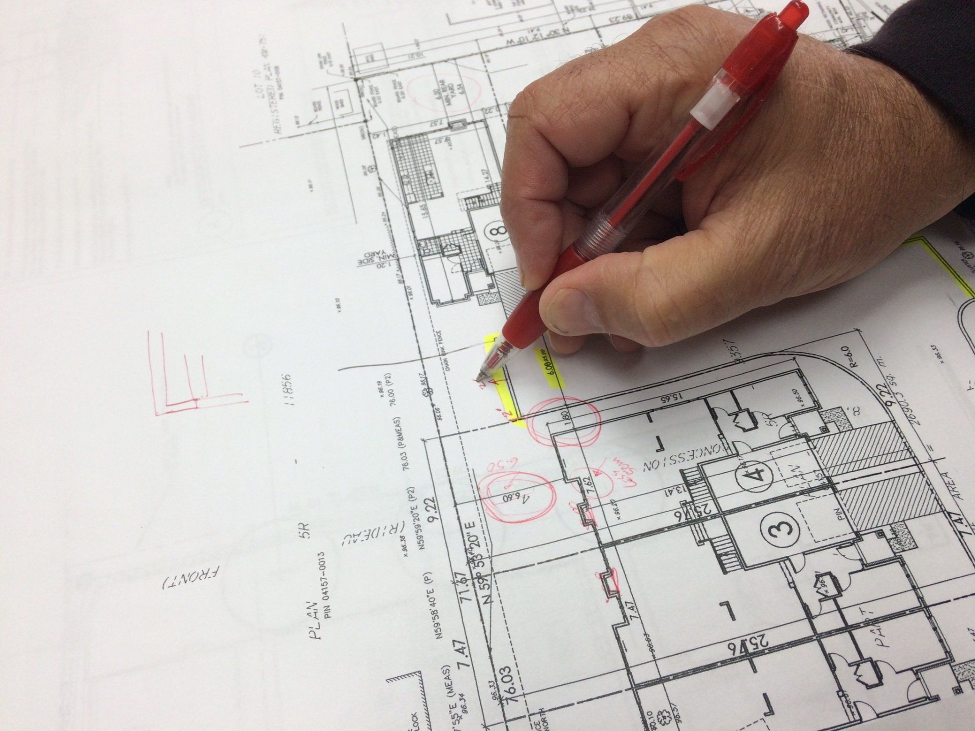 Une main tenant un stylo rouge marque un plan de construction avec un surligneur jaune.