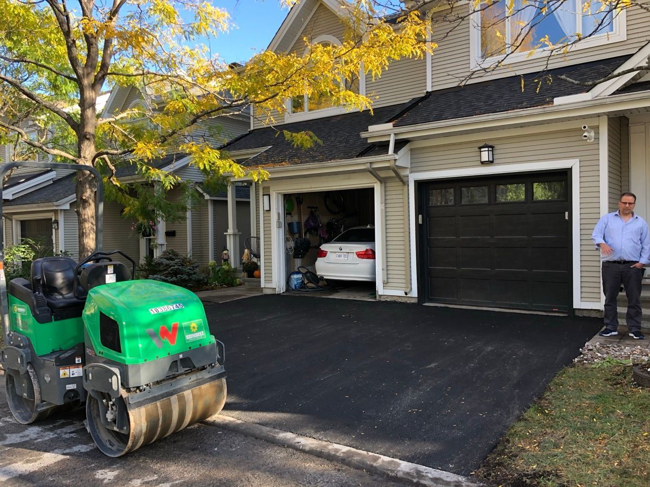 Rouleau compresseur pour asphalte compactant une nouvelle allée ; un homme regarde près du garage, dans un cadre suburbain.